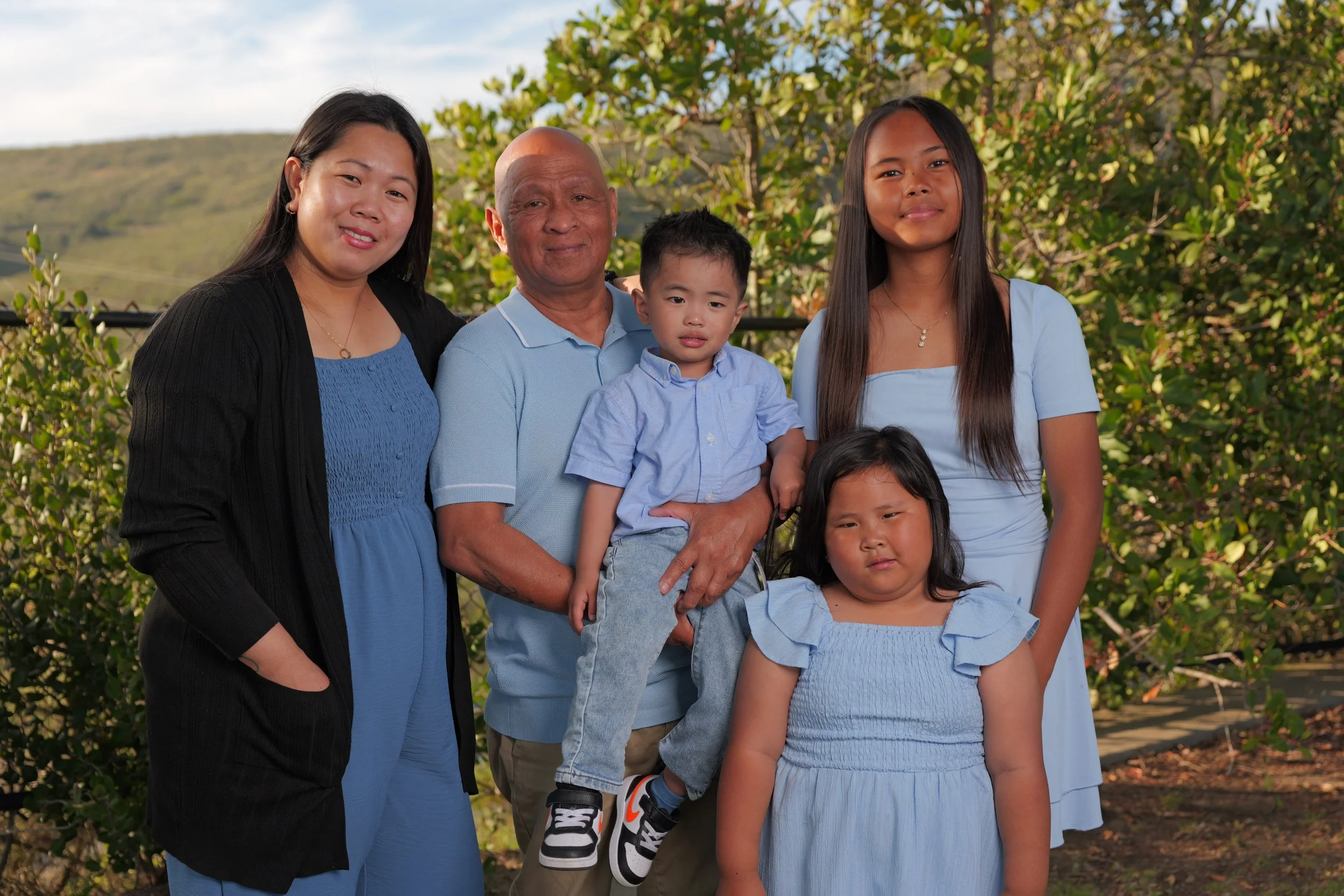 Group of five people, including two adults and three children, standing outdoors in front of green foliage and a hill, posing for a family photo.