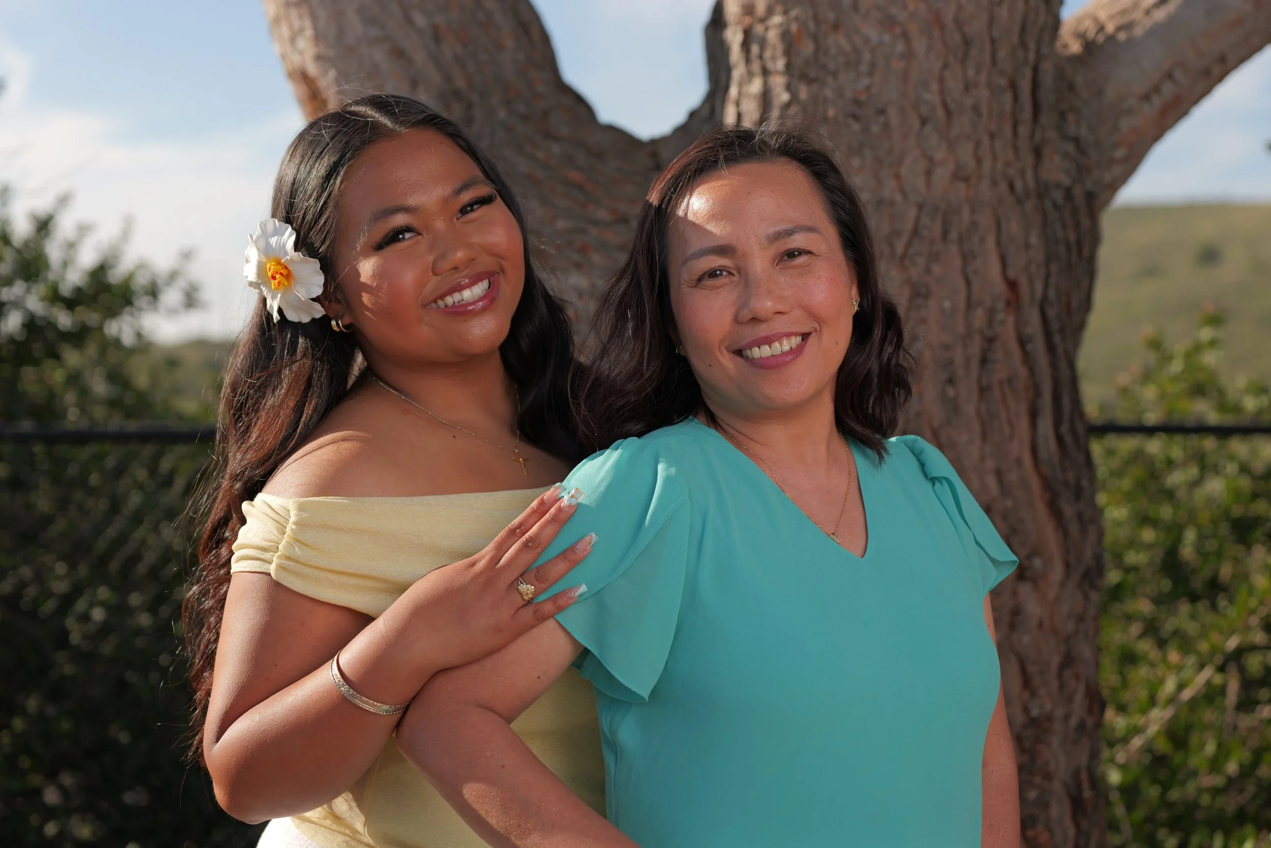 Two women smiling outdoors in front of a tree, one with a yellow off-shoulder dress and a flower in her hair, the other in a light blue top.