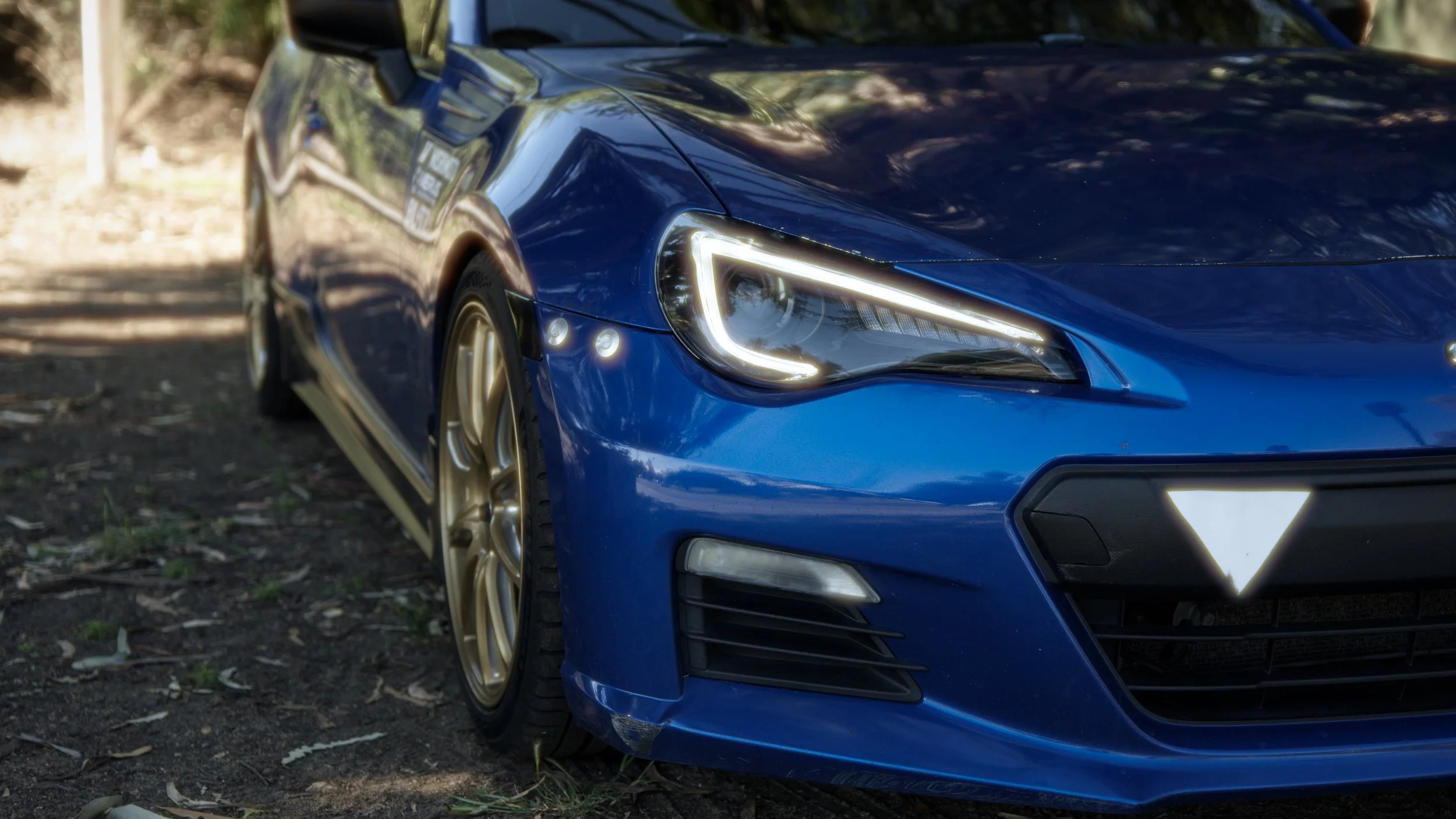 Close-up of the front right side of a blue sports car with gold wheels parked on dirt ground with trees in the background.
