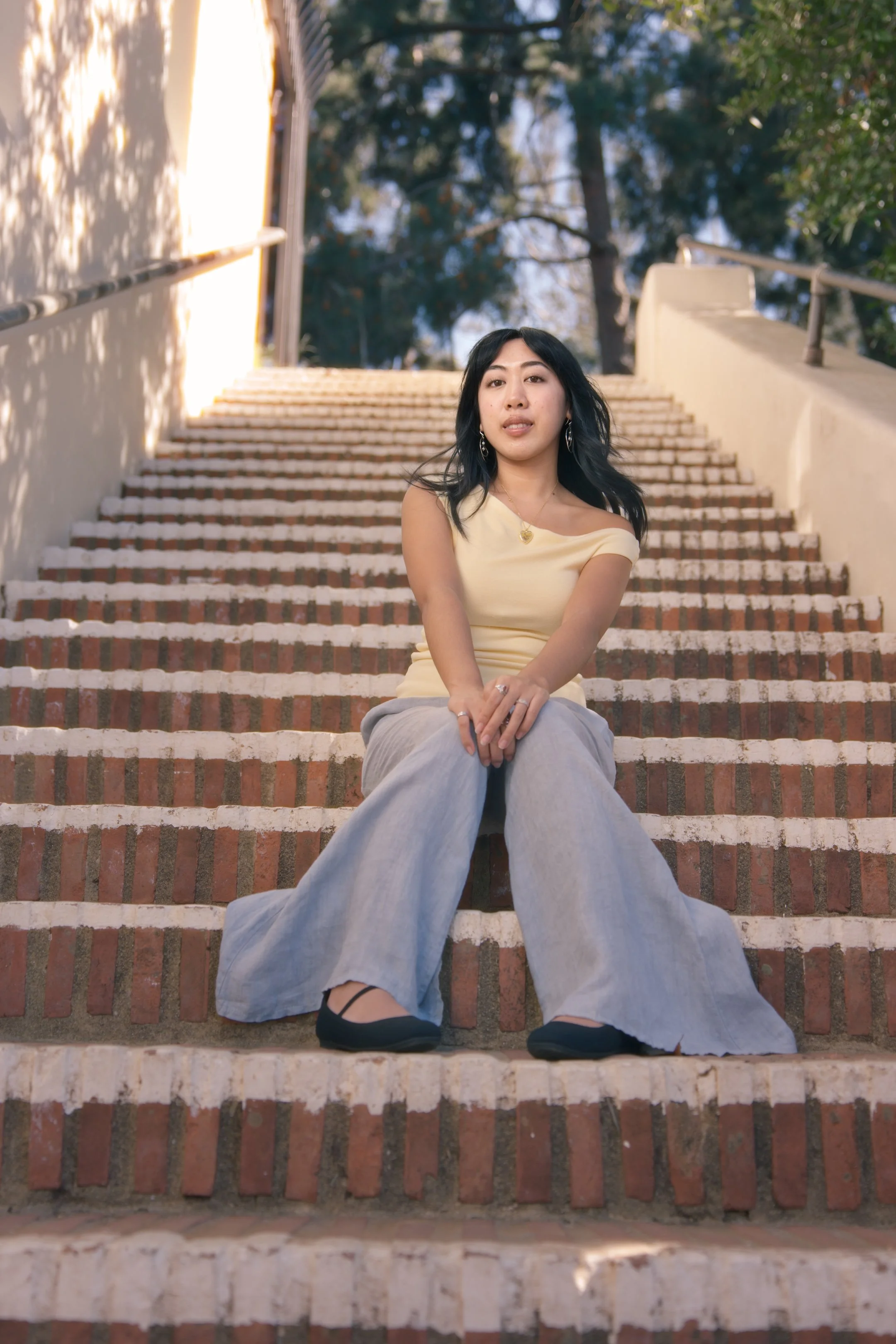 A young woman with black hair sitting on brick steps outdoors, wearing a light yellow top and loose gray pants, with trees in the background.