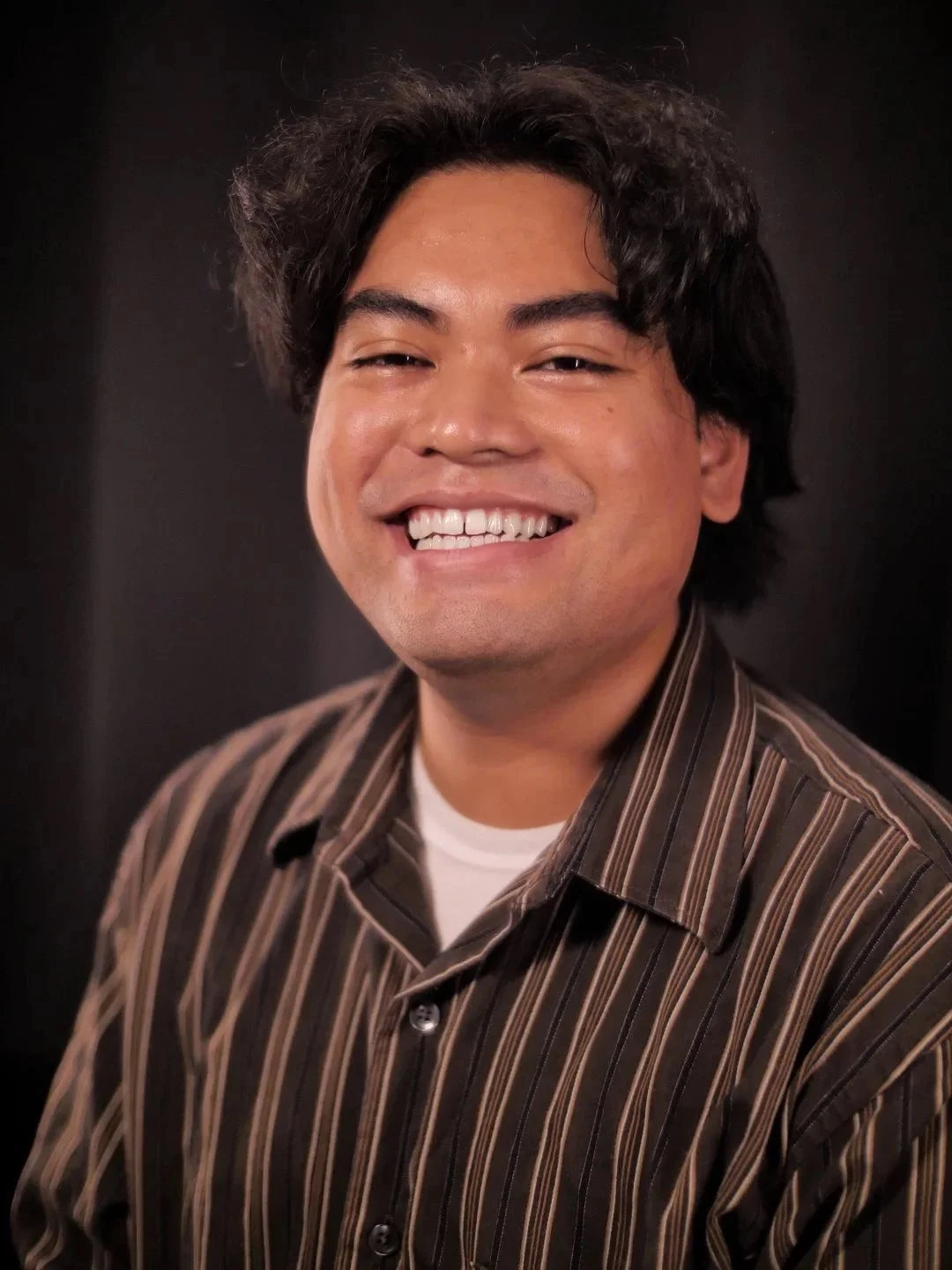 A smiling man with dark, wavy hair wearing a brown striped shirt against a dark background.