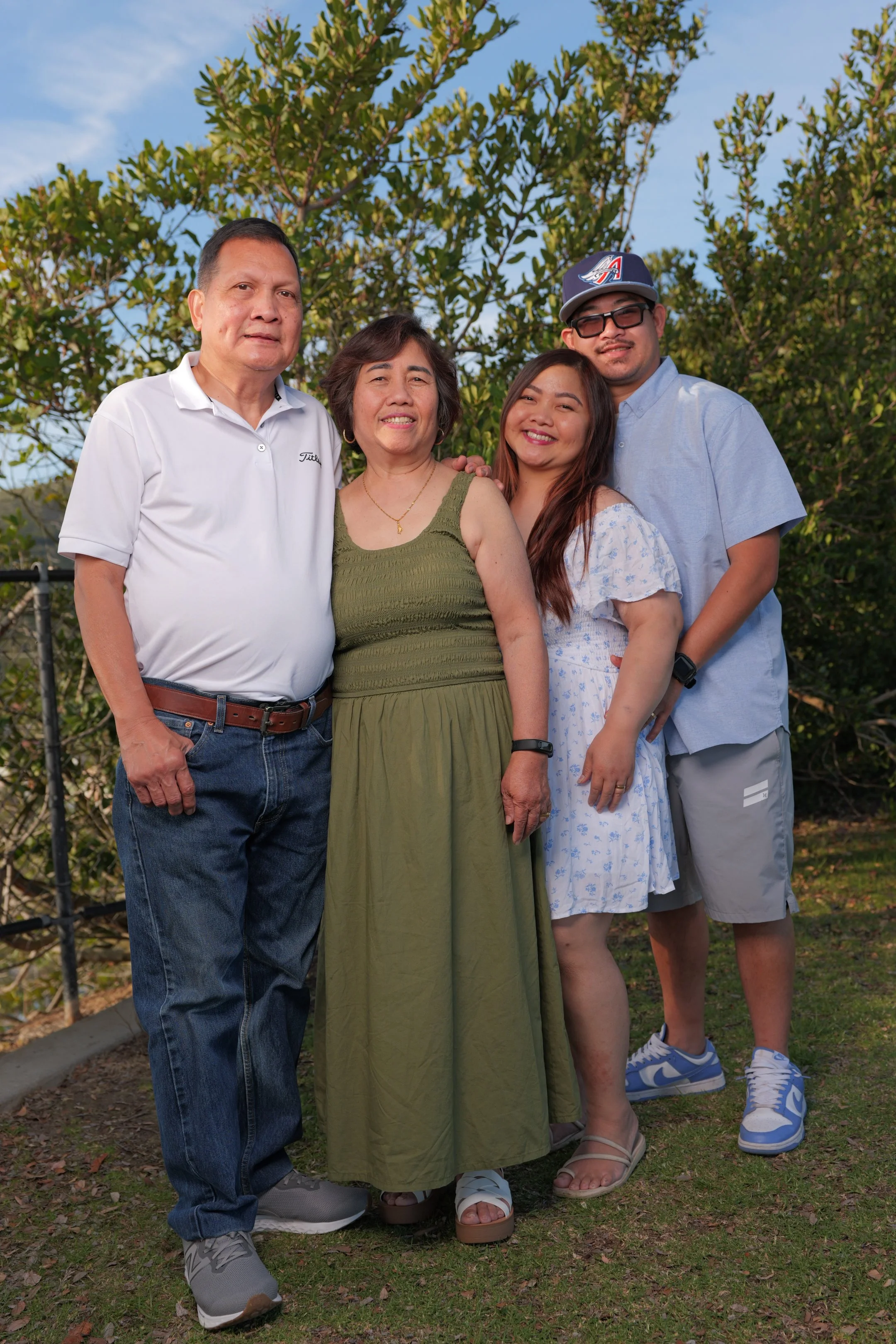 A family of four standing outdoors on grass with trees in the background, smiling at the camera, during the daytime.