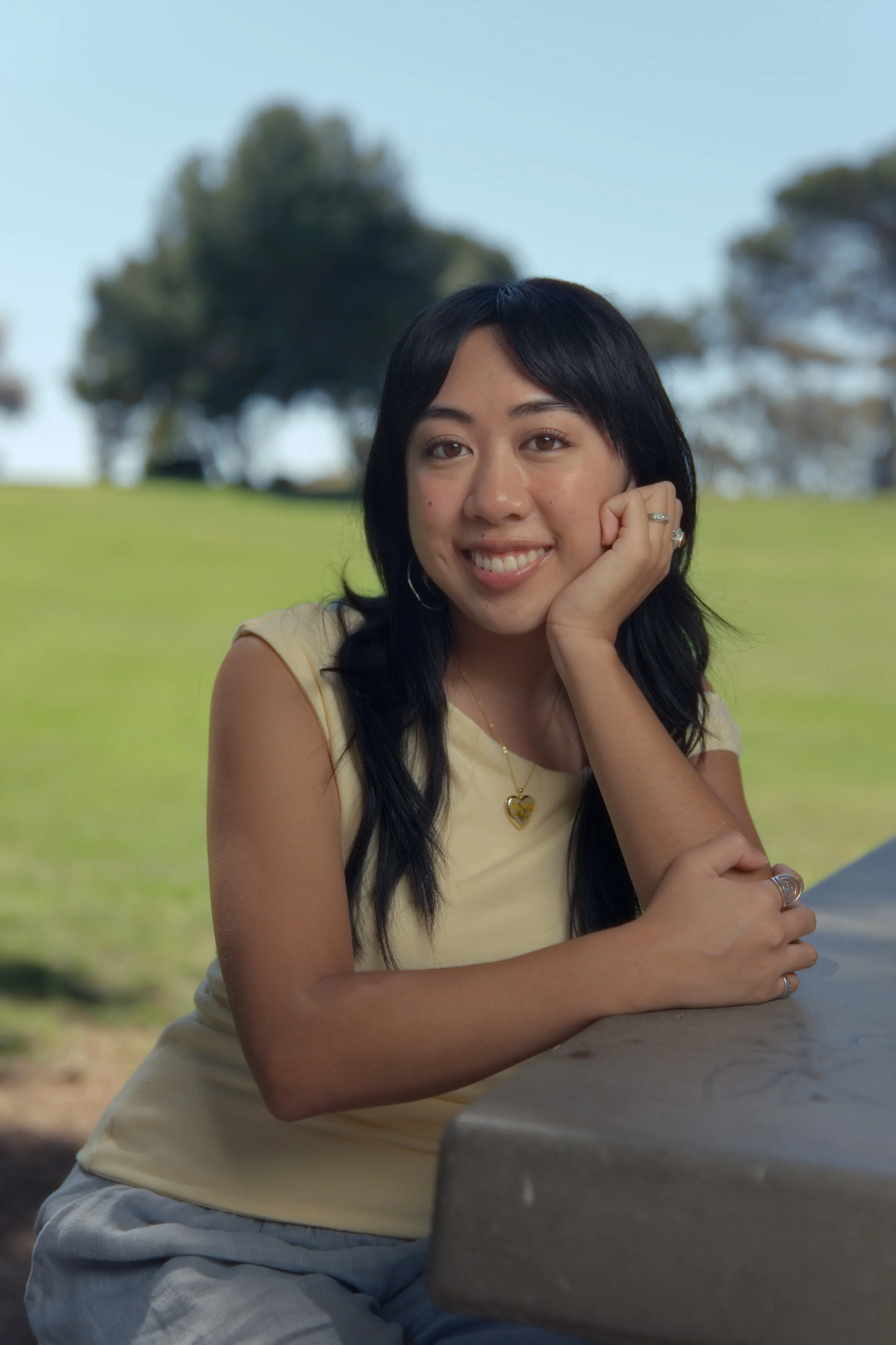 A young woman with dark hair, smiling, resting her head on her hand, sitting outdoors at a park table with grassy area and trees in the background.