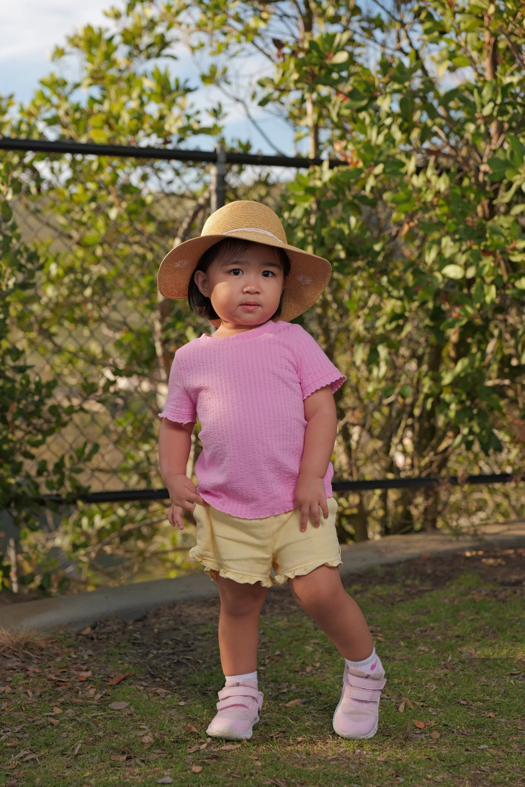 Young girl wearing a pink shirt, yellow shorts, white shoes, and a straw hat standing outdoors on grass with green bushes and trees in the background.