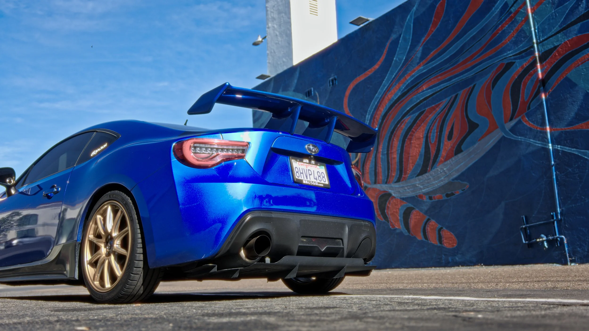 Blue sports car with gold wheels and a large rear spoiler parked in front of mural art on a building wall, under a clear blue sky.