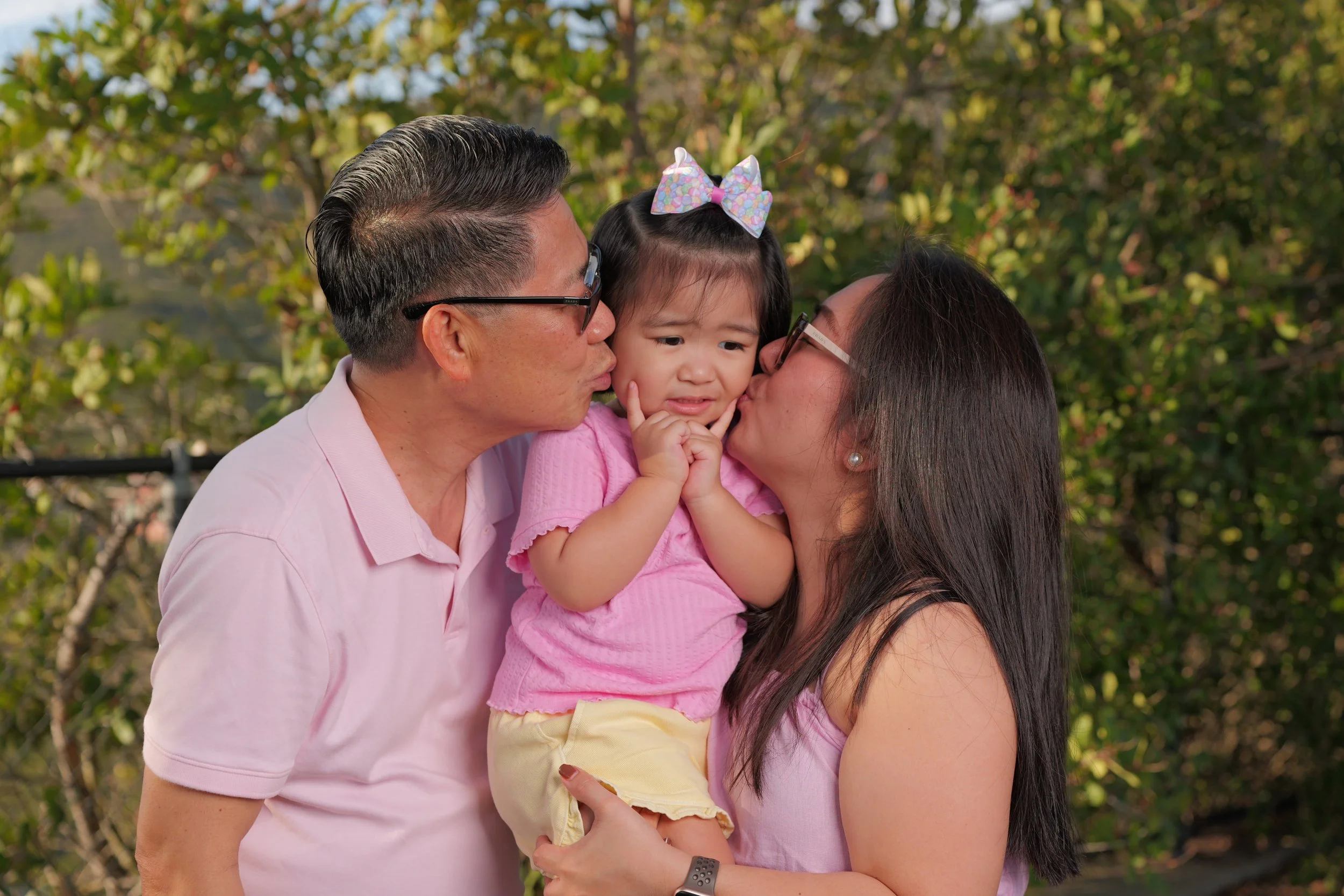 A young girl wearing a pink shirt and a colorful bow in her hair is being kissed on both cheeks by her parents, who are wearing matching pink shirts. The family is outdoors with green trees in the background, and the girl appears slightly unsure.