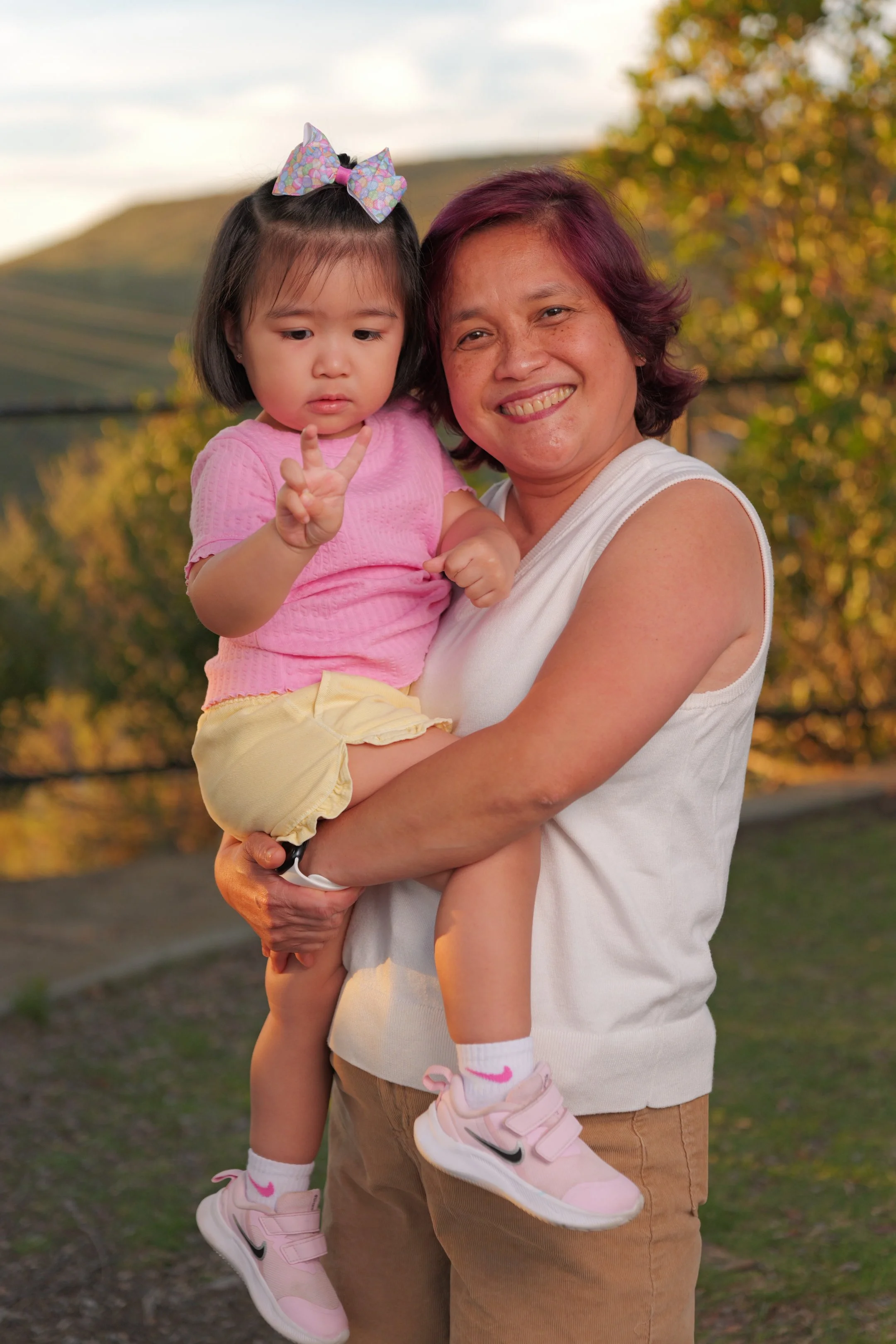 A woman smiling while holding a young girl with a peace sign in an outdoor park setting during sunset.