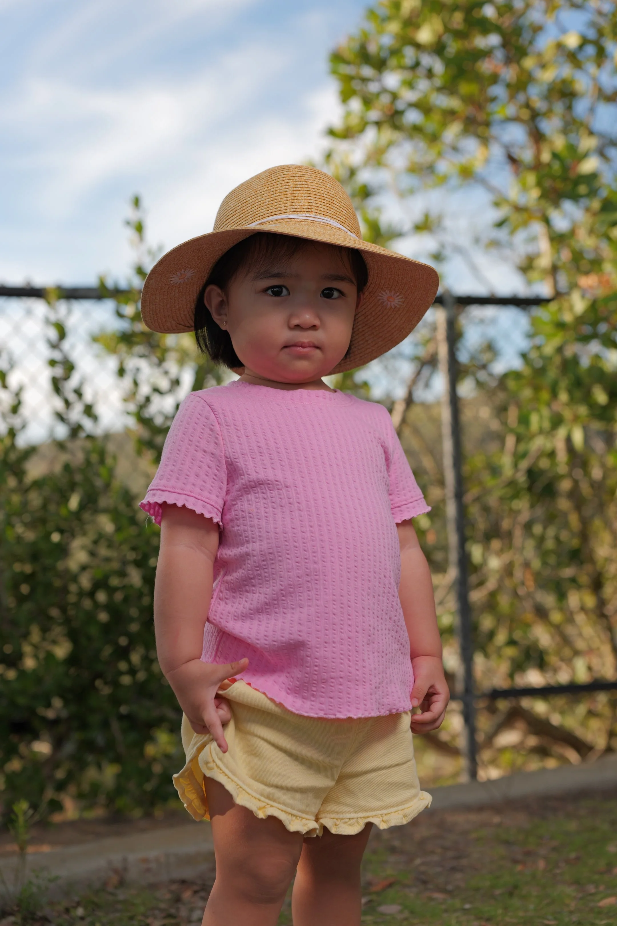 A young girl wearing a pink textured shirt, yellow shorts, and a large straw hat, standing outdoors near trees with a cloudy sky background.