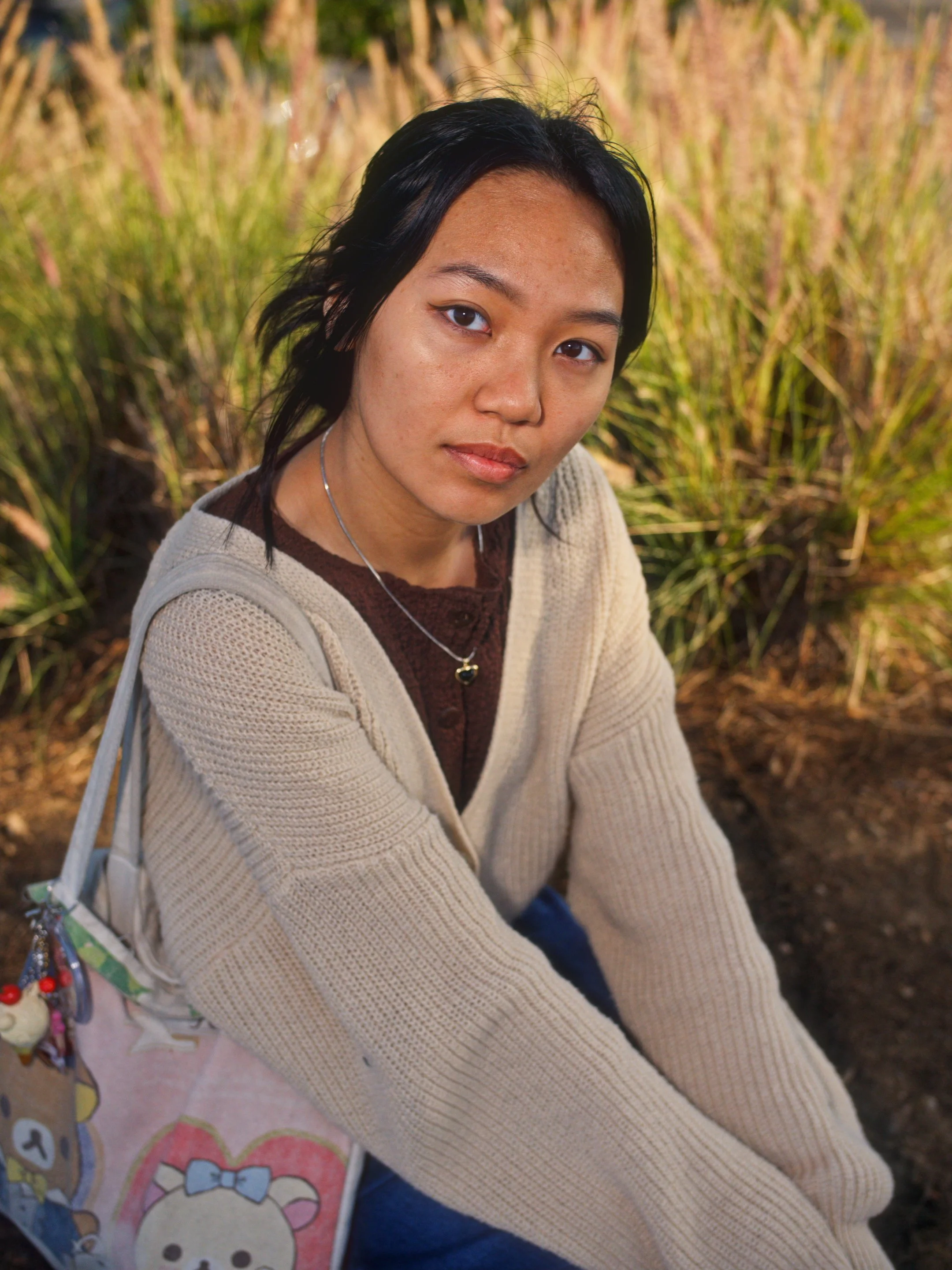 A young woman with black hair and tan skin sitting outdoors in a grassy area, wearing a beige cardigan, a dark shirt, and a necklace, with a colorful bag on her side.