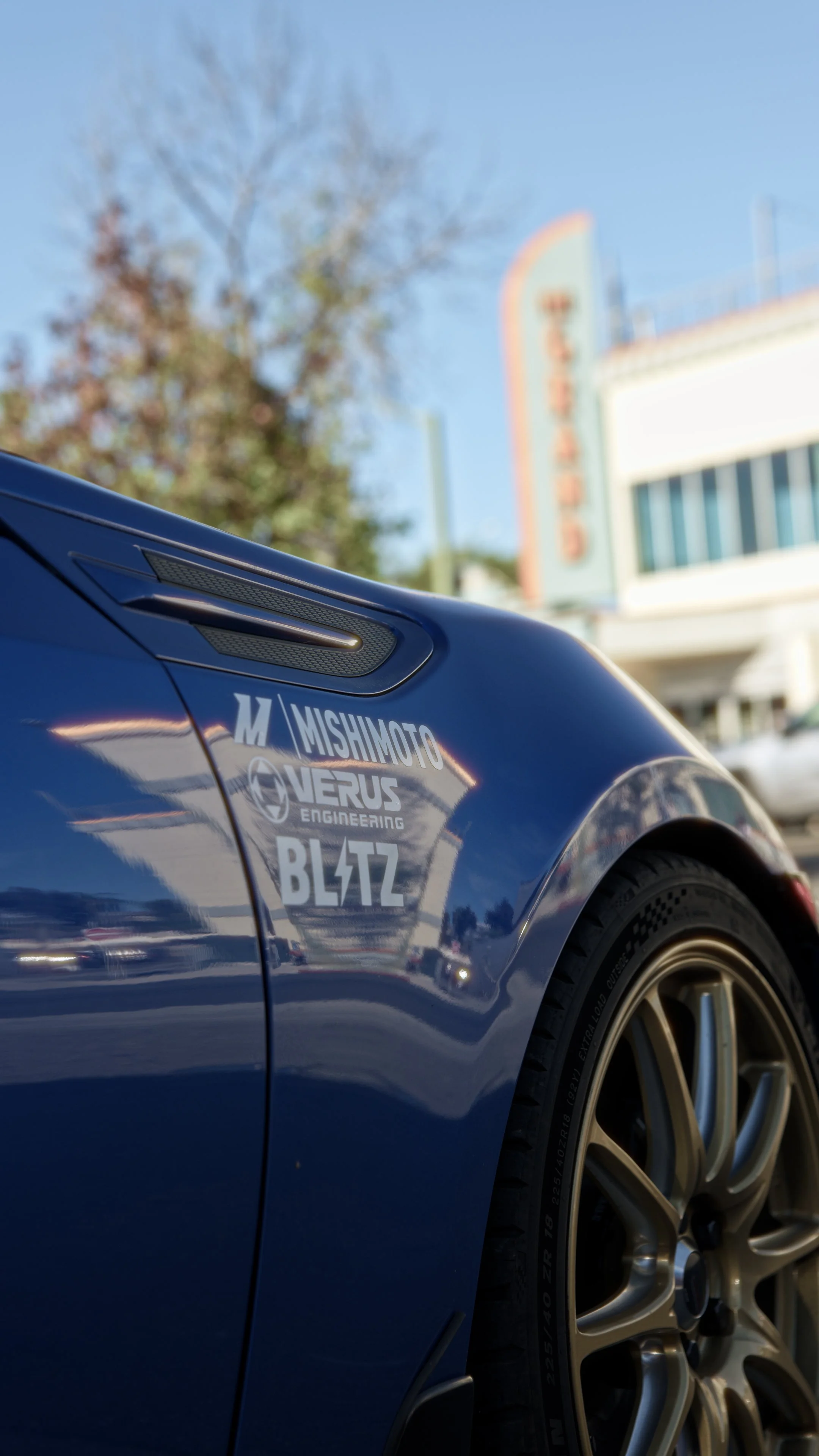 Close-up of a blue sports car with racing and engineering decals on its side, shown outdoors against a clear sky with a tree and building in the background.