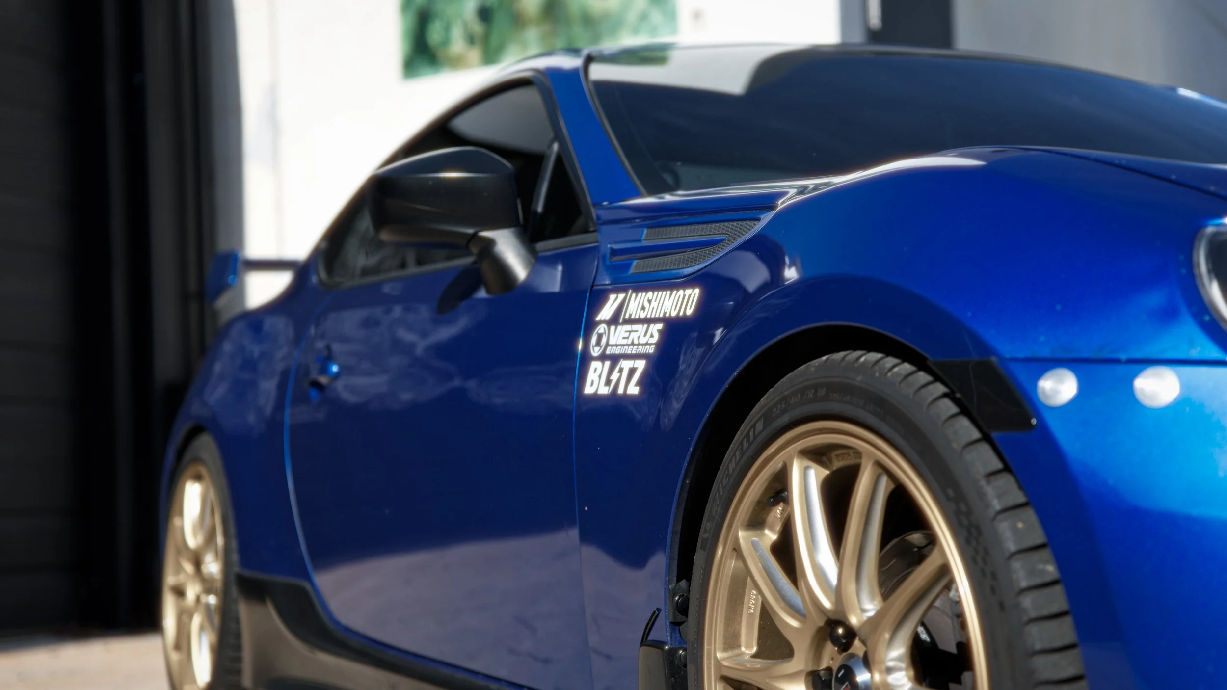 Close-up of a blue sports car with gold wheels, featuring decals including Mishimoto, Verus Engineering, and Blitz.