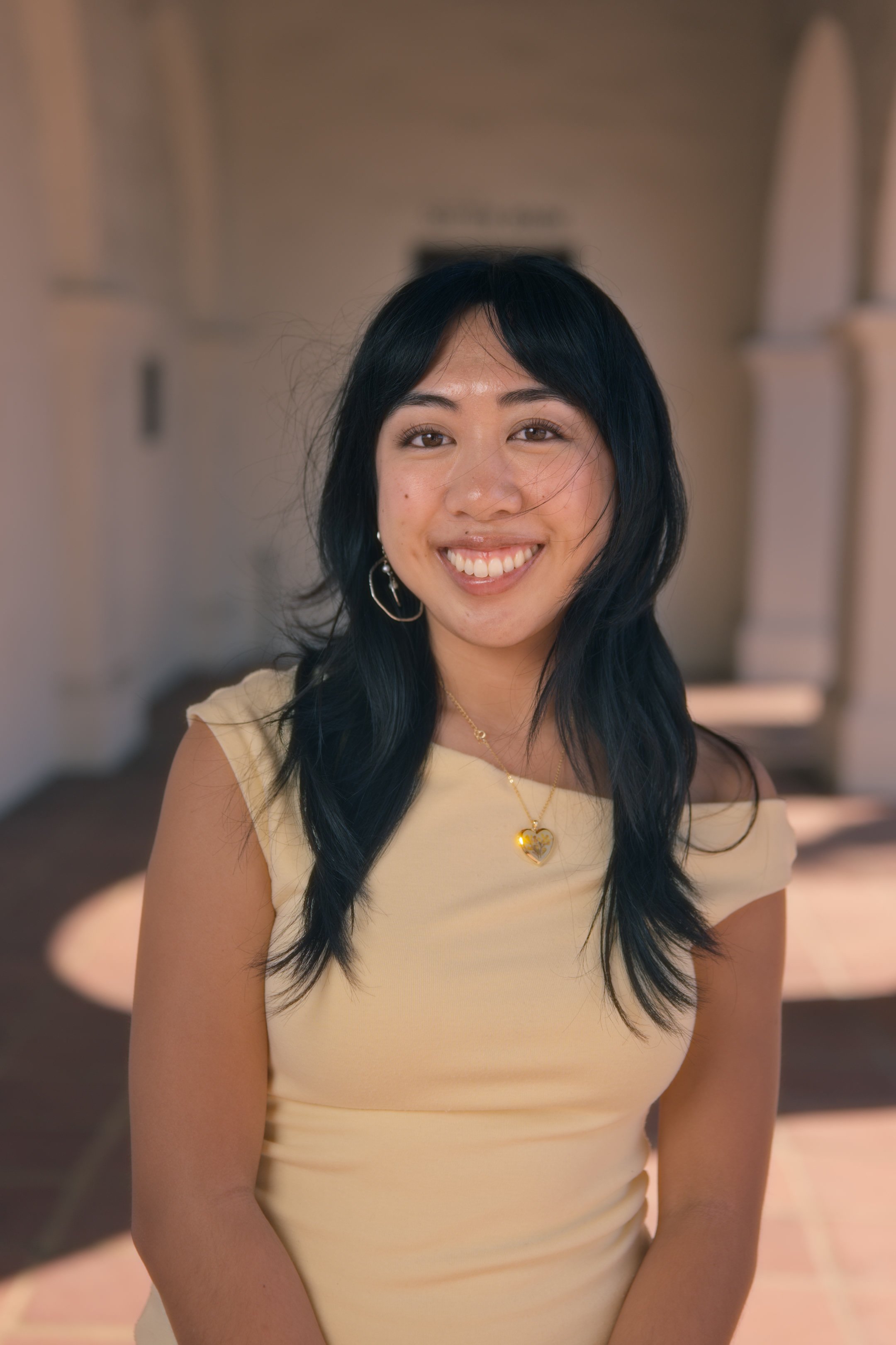 A woman with black hair styled in waves, smiling and wearing gold jewelry including hoop earrings, a heart-shaped pendant necklace, and a delicate chain. She is dressed in a light yellow sleeveless top and is standing indoors with a blurred backgroun