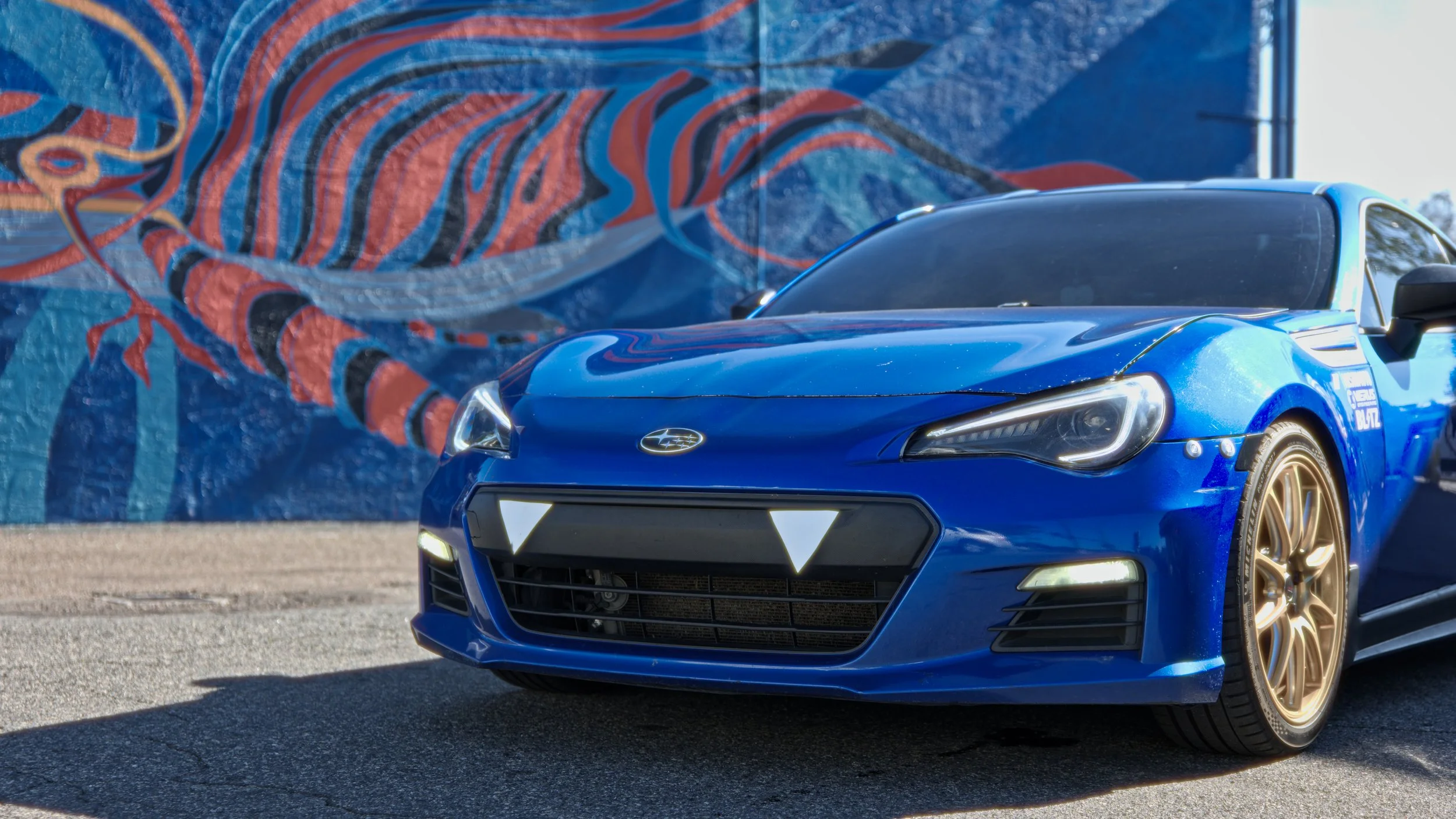 Blue sports car with gold rims parked in front of a mural with abstract colors and shapes.