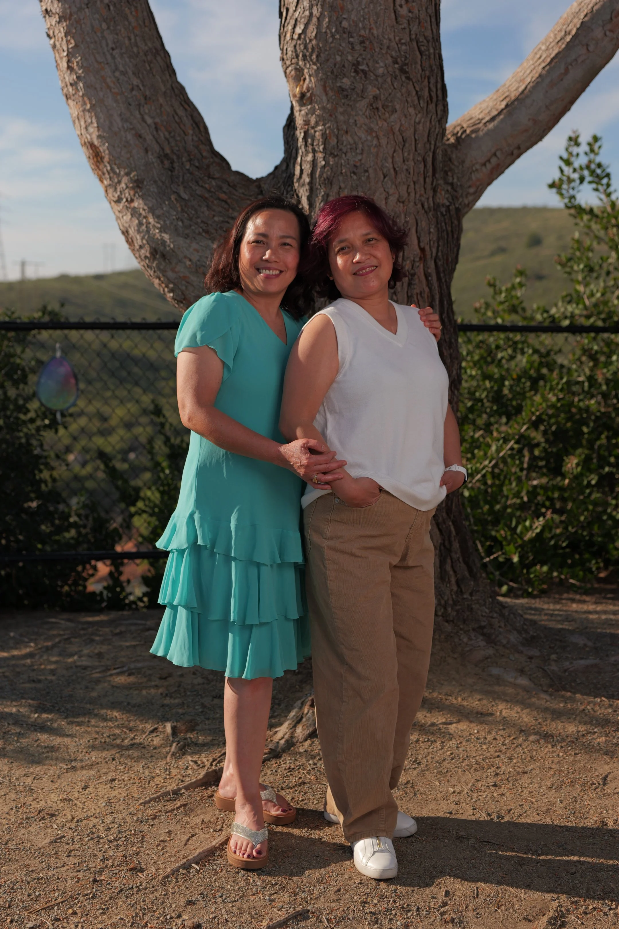 Two women standing outdoors in front of a large tree, smiling, with a chain-link fence and green hills in the background.