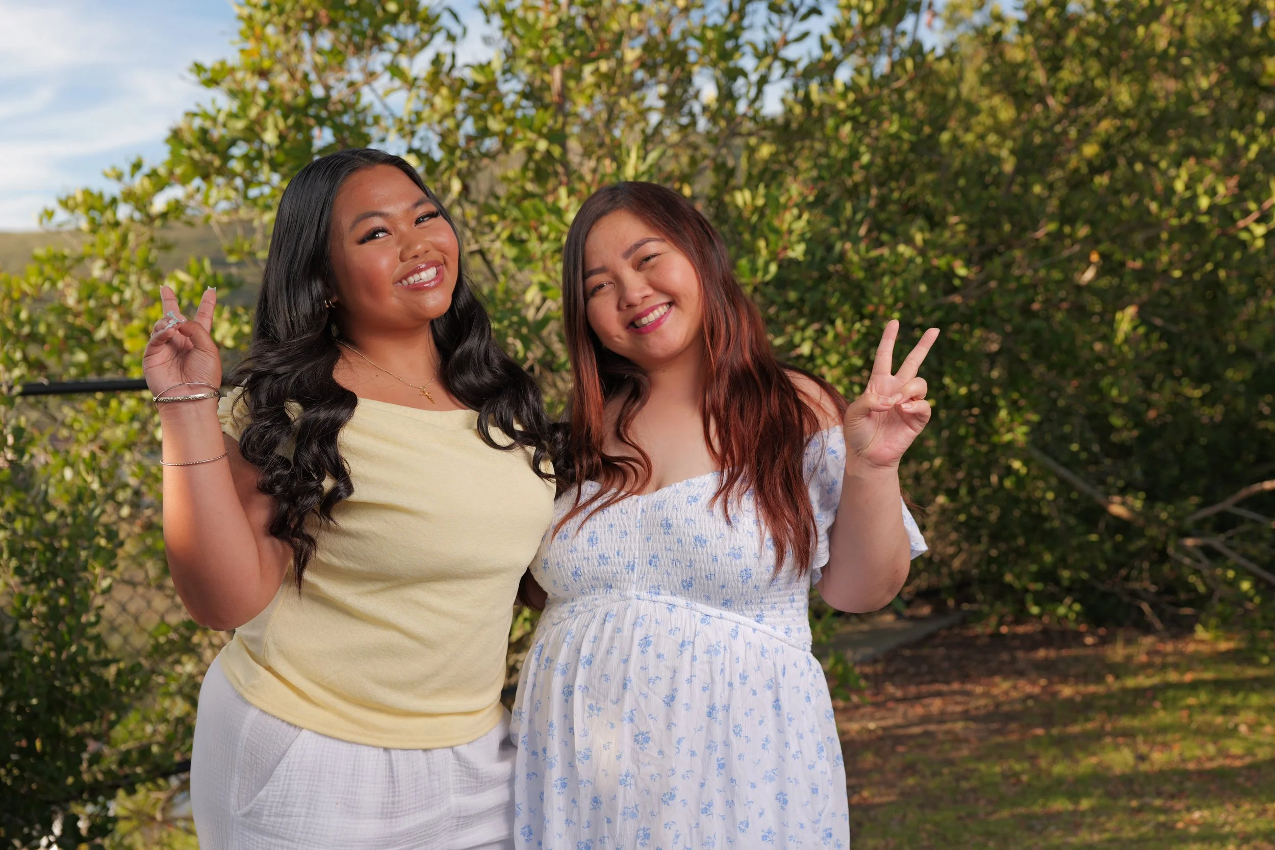 Two young women smiling and making peace signs with their hands outdoors in front of greenery.