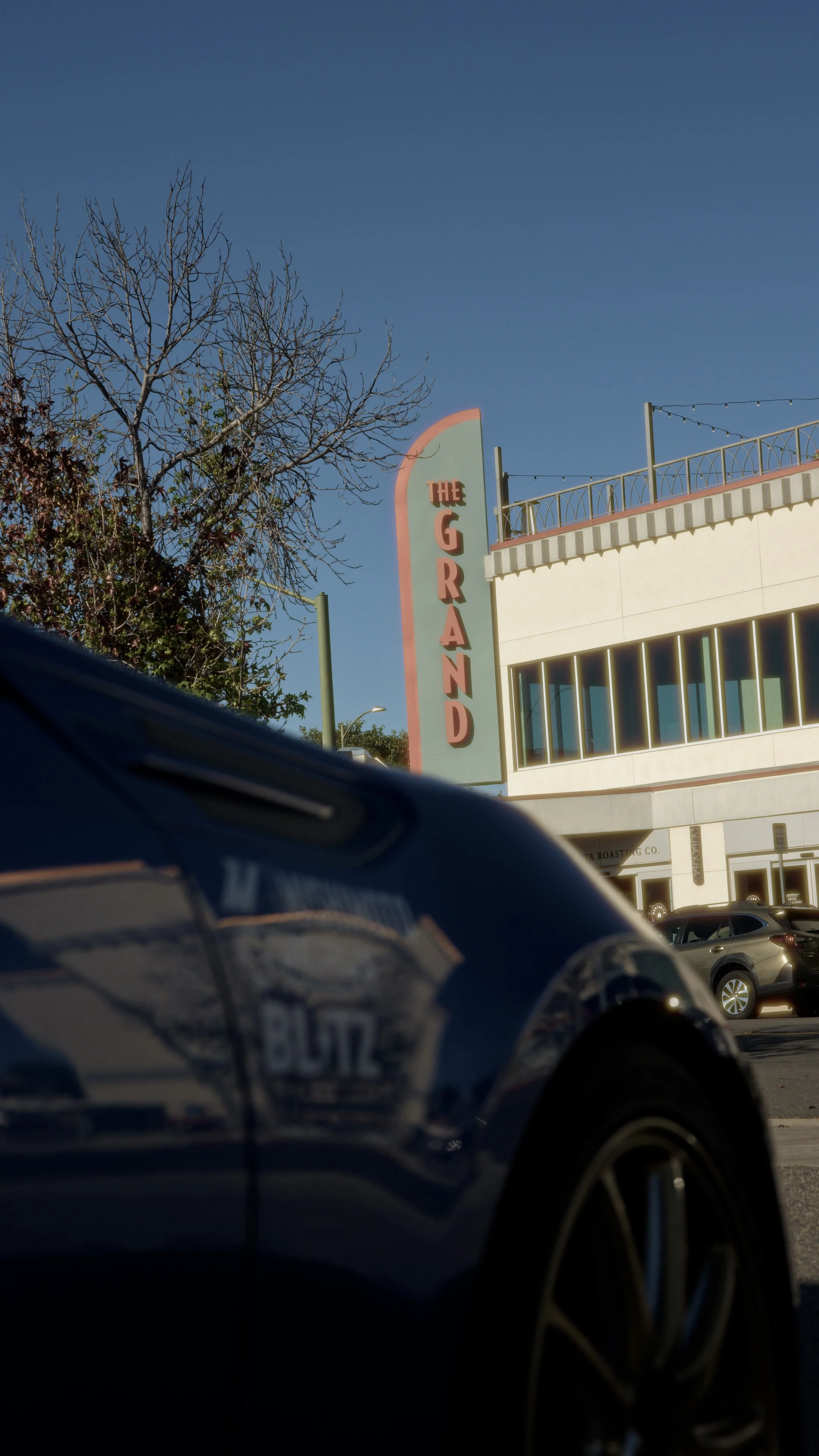 Partially visible parked car in the foreground, storefront with sign 'The Grand' and large windows, leafless tree, and a clear blue sky.