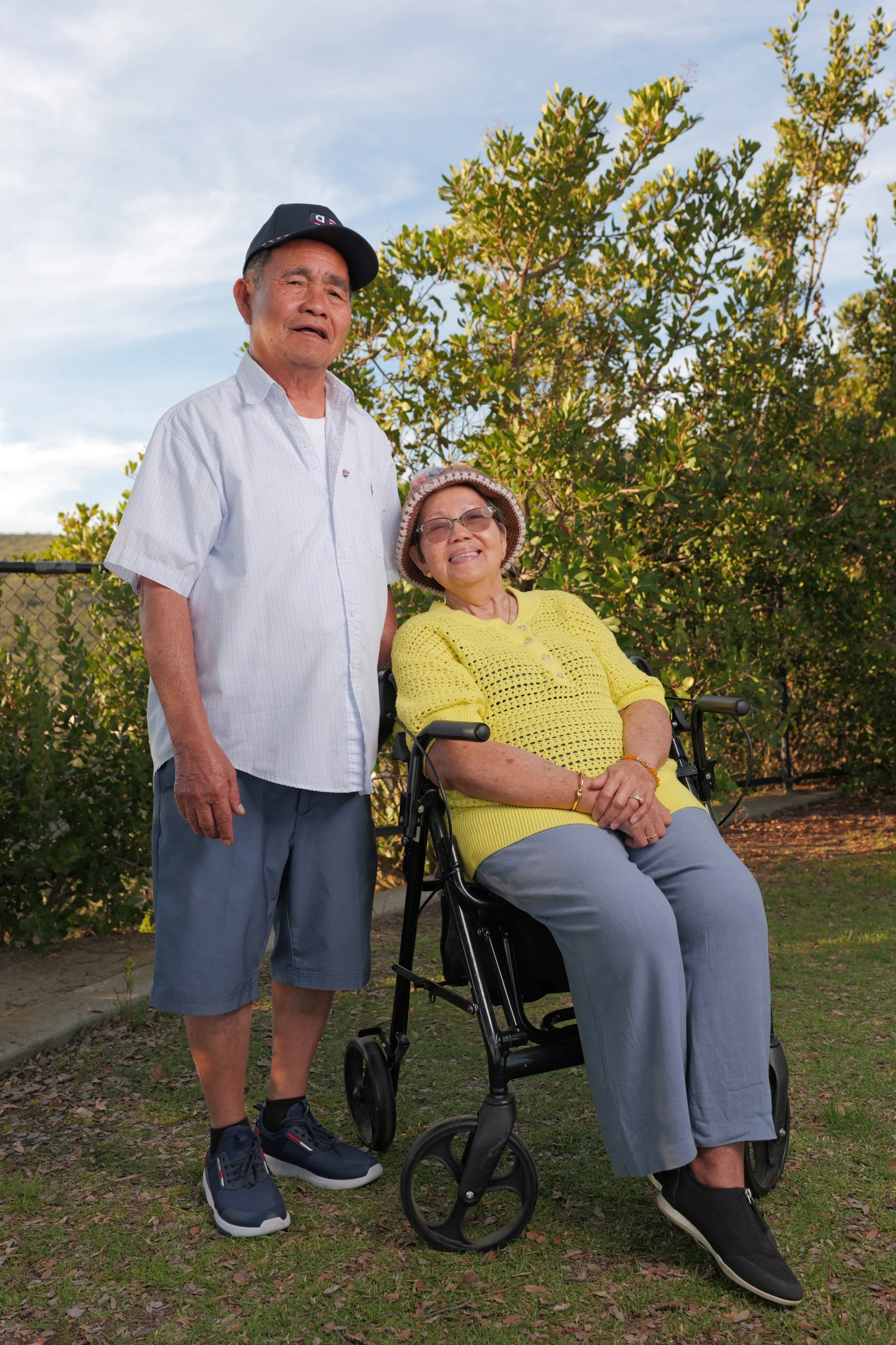 An elderly woman in a wheelchair outdoors, smiling, with an elderly man standing beside her, on a sunny day with trees and a fence in the background.