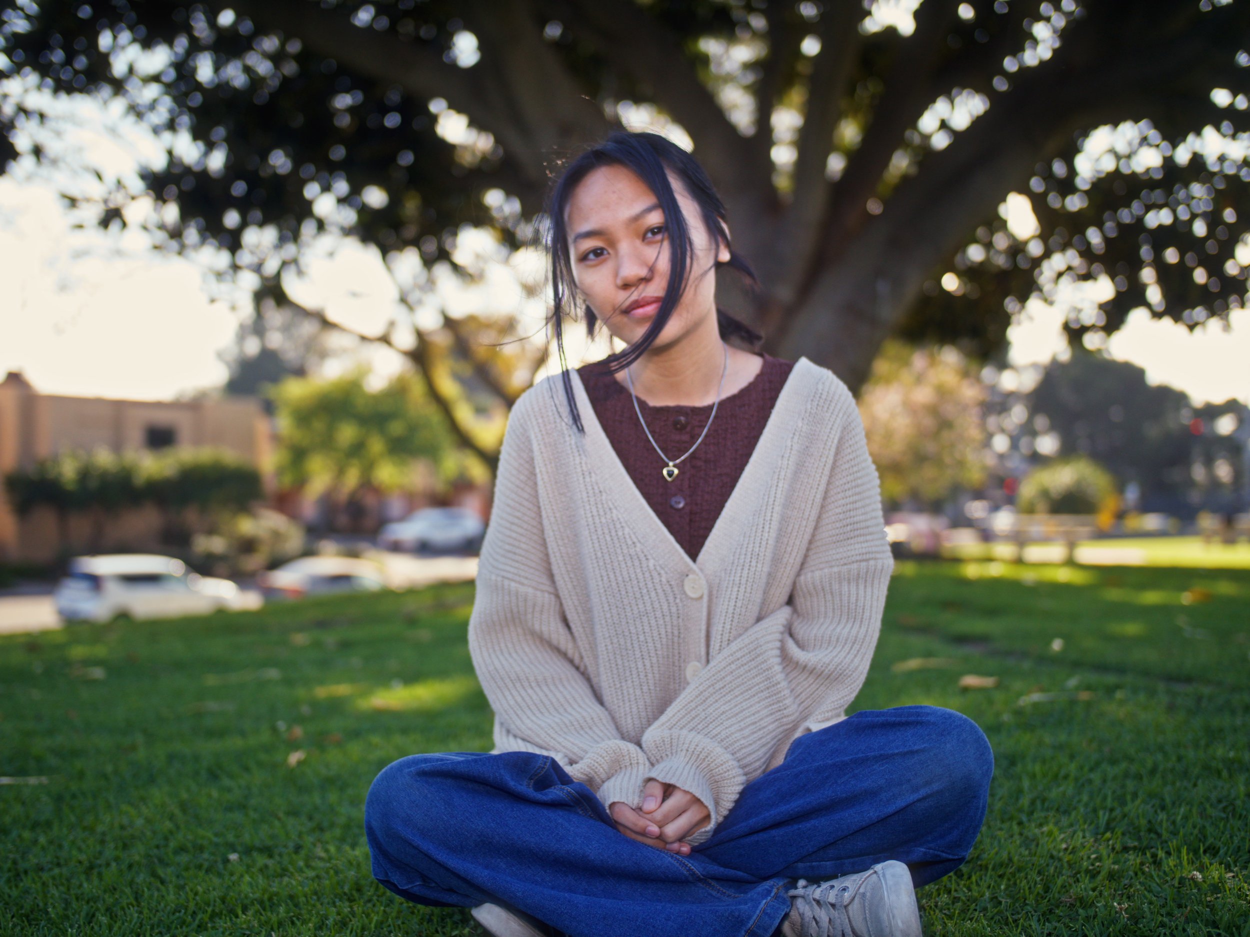 A young woman sitting cross-legged on grass under a large tree in a park, with cars and buildings in the background, during the daytime.