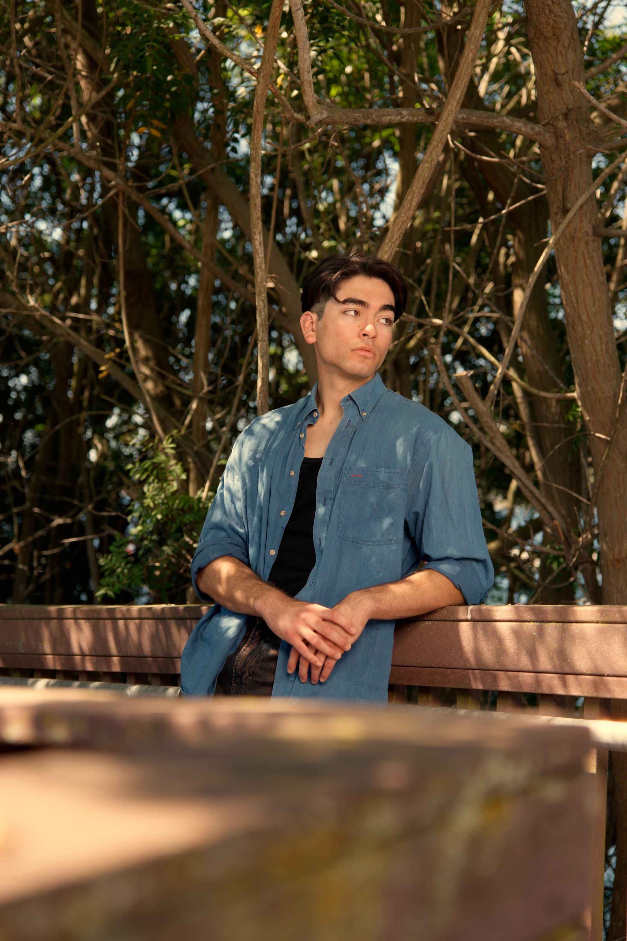A young man with dark hair, wearing a blue denim shirt over a black t-shirt, leaning on a wooden railing outdoors, with trees and dense foliage in the background.