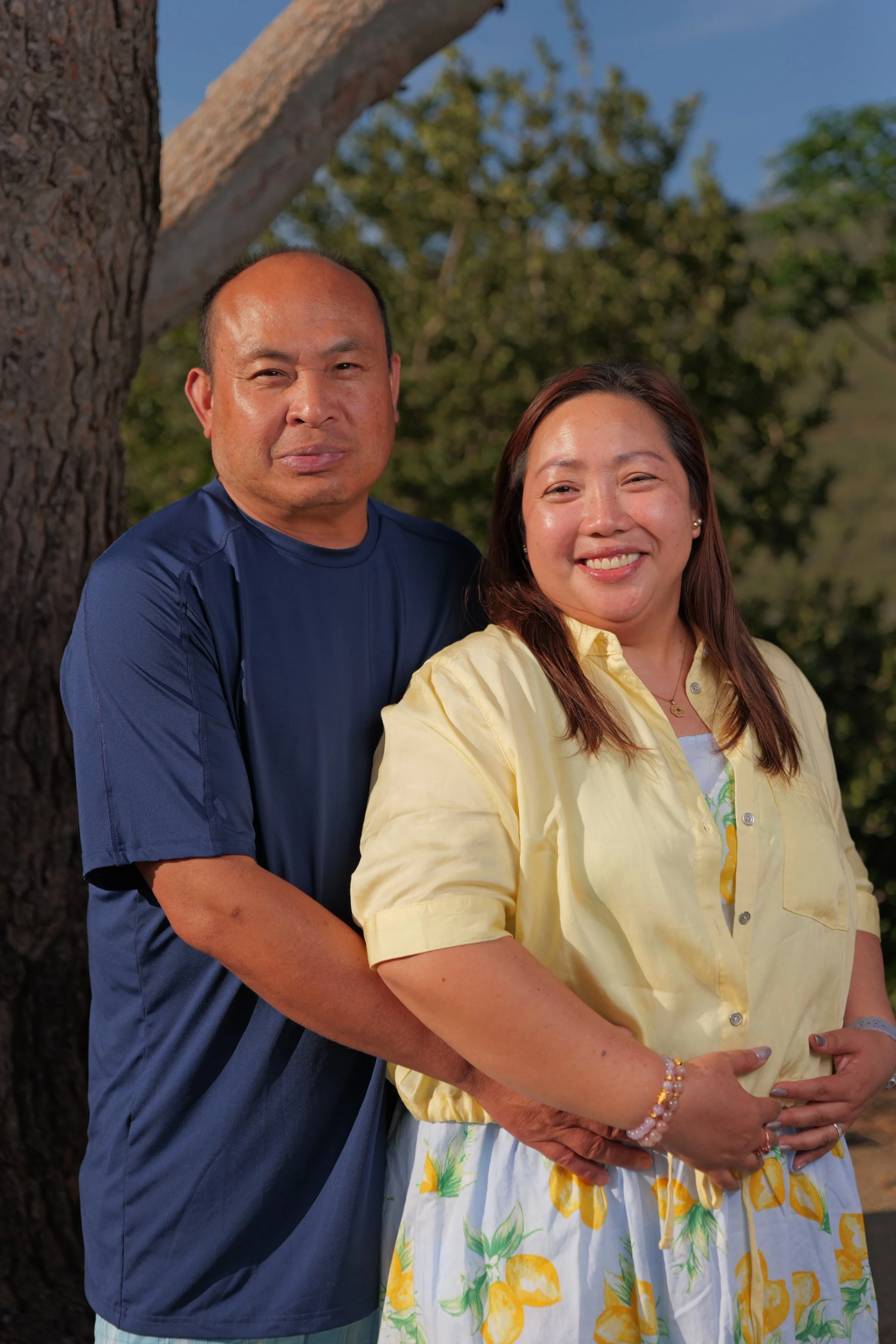 A man and woman standing outdoors near a tree, with sunlight illuminating their faces and a blurred natural background of green trees and blue sky.