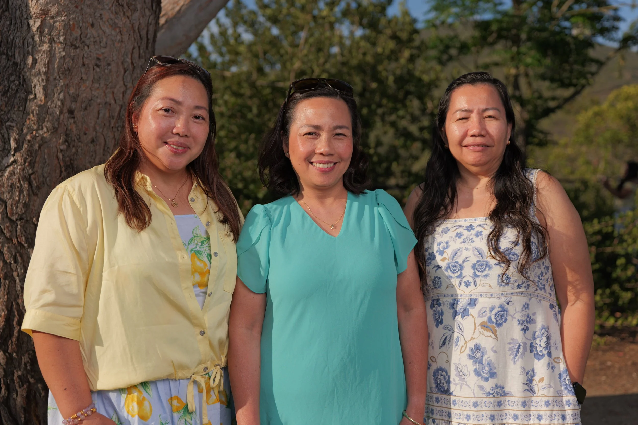 Three women standing outdoors in front of a tree with green foliage, smiling at the camera.