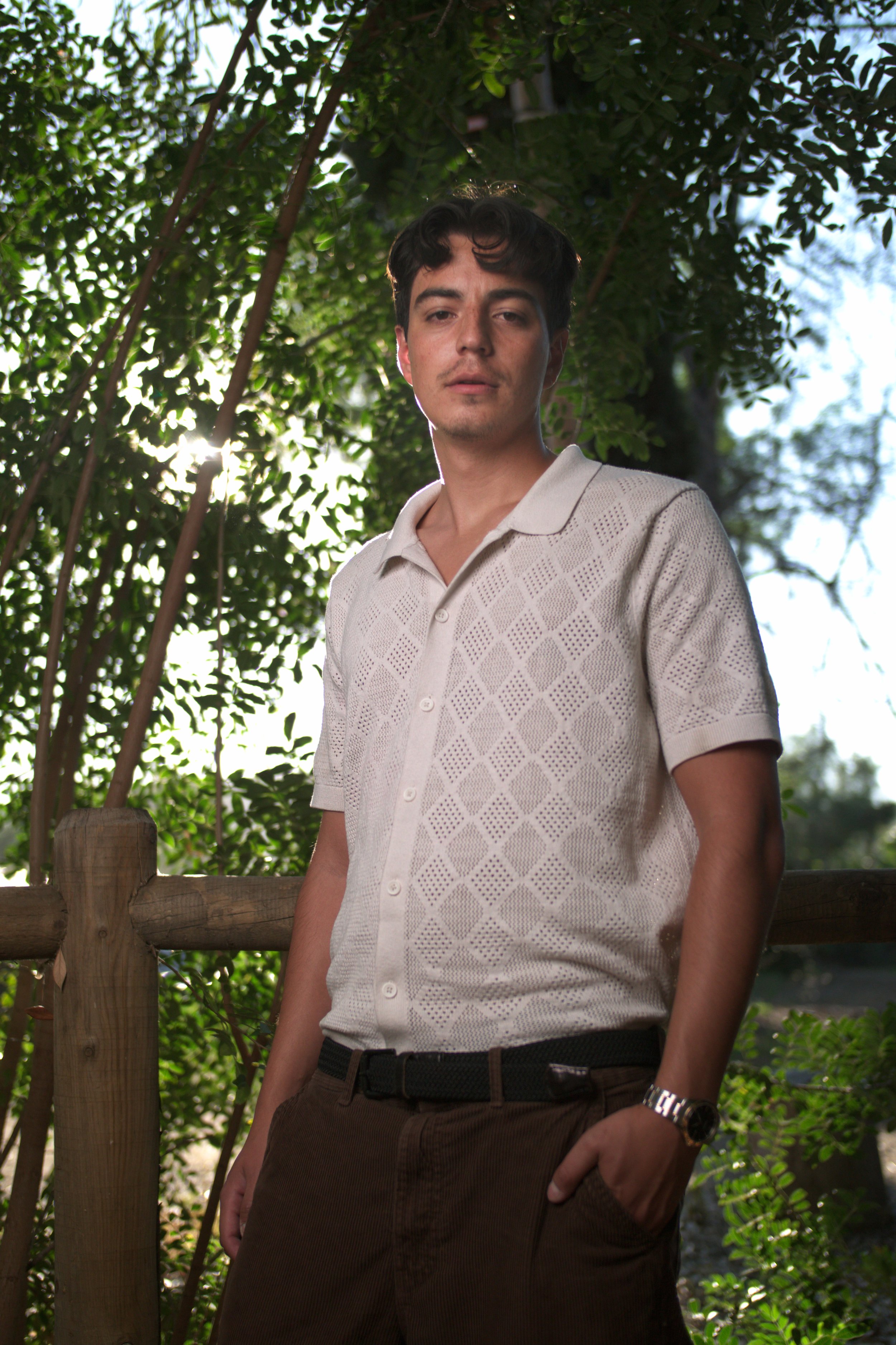 A young man stands outdoors near a wooden railing, wearing a cream-colored short-sleeve shirt and brown pants, with lush green foliage and sunlight in the background.