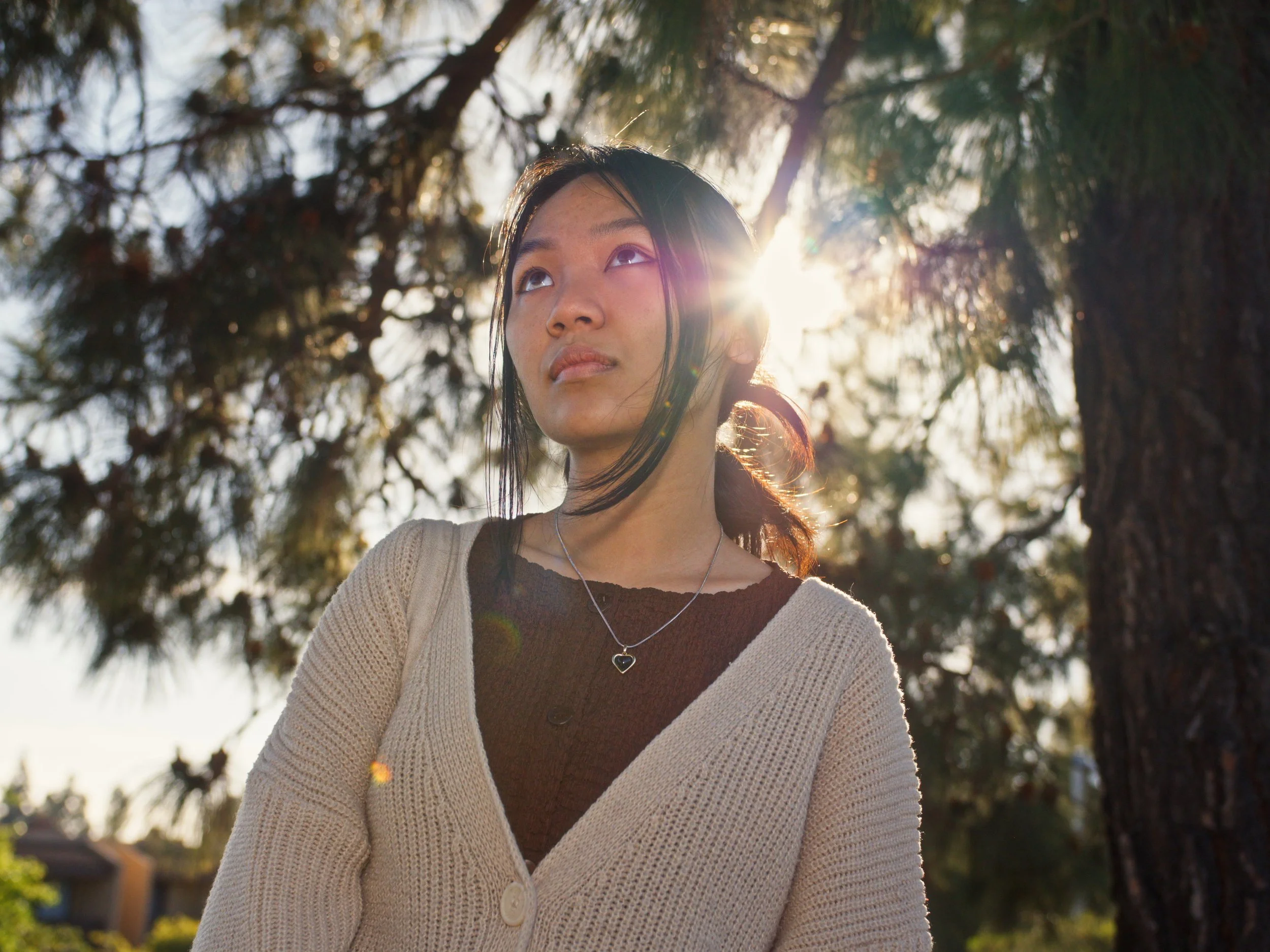 A woman with dark hair looking up outdoors with sunlight behind her, trees in the background.
