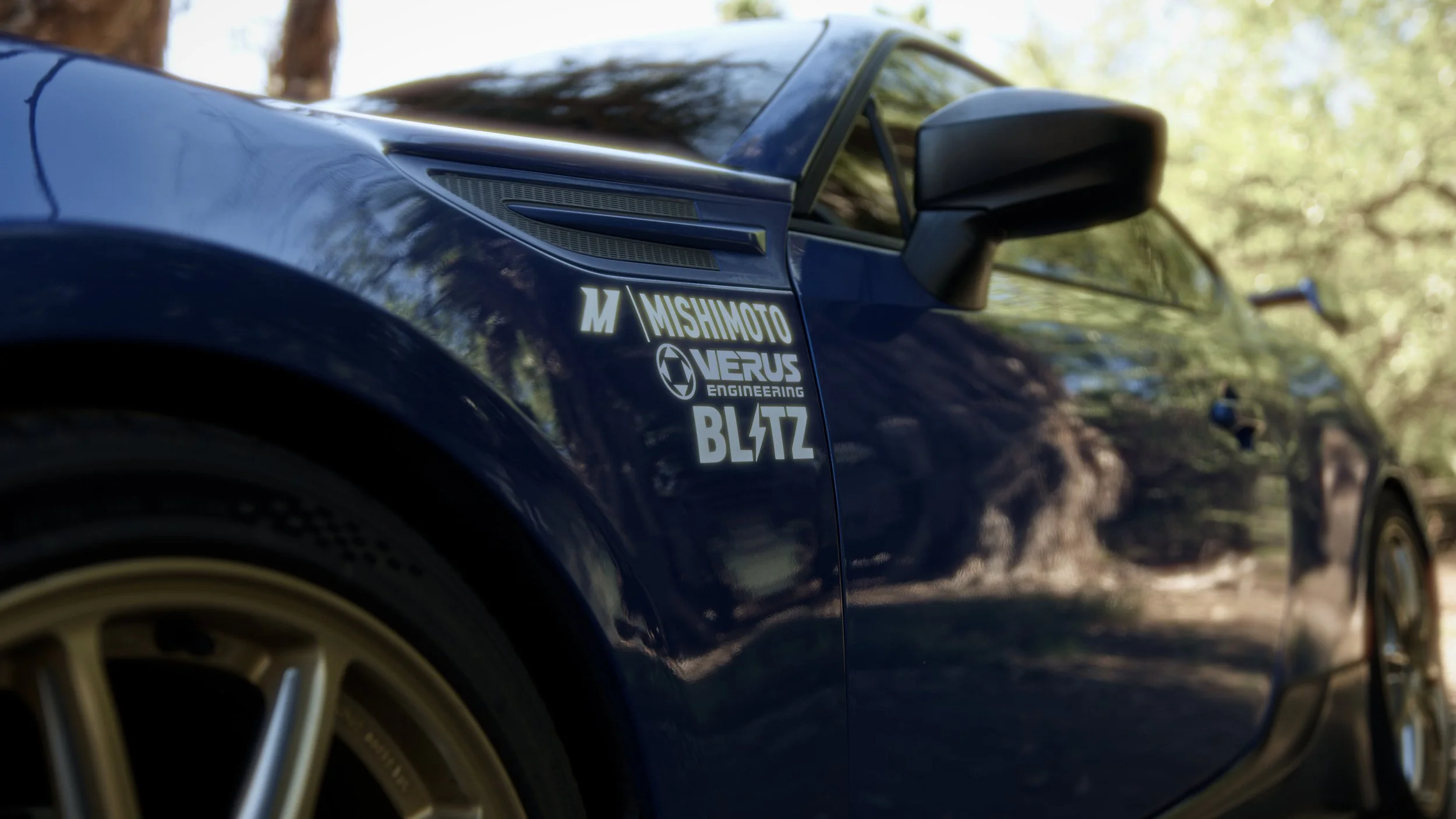Close-up of a blue sports car with sponsor stickers, including Mishimoto, Verus Engineering, and Blitz, on the side near the front wheel, parked outdoors among trees with reflections on the car's surface.