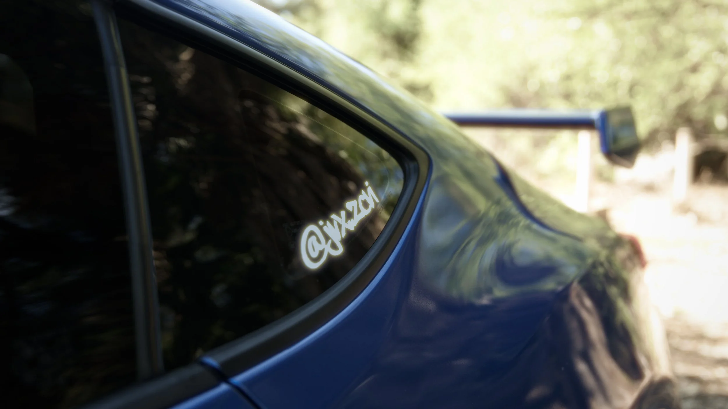 Close-up of a dark-colored car with a stick-on decal of the Twitter handle '@jwxzgh' on the rear side window, with a blurred outdoor background.