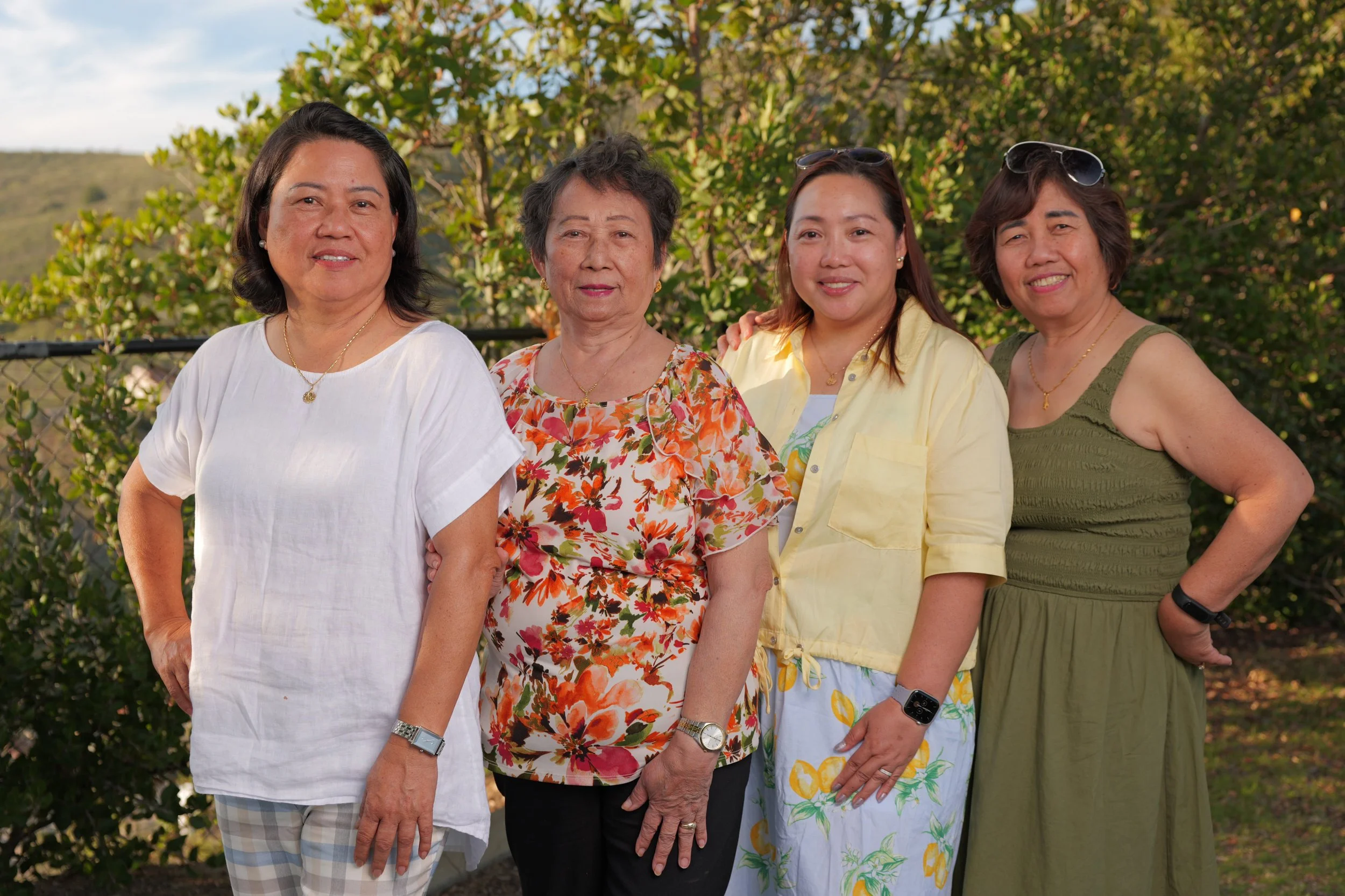Four women standing outdoors in front of green foliage, smiling at the camera.