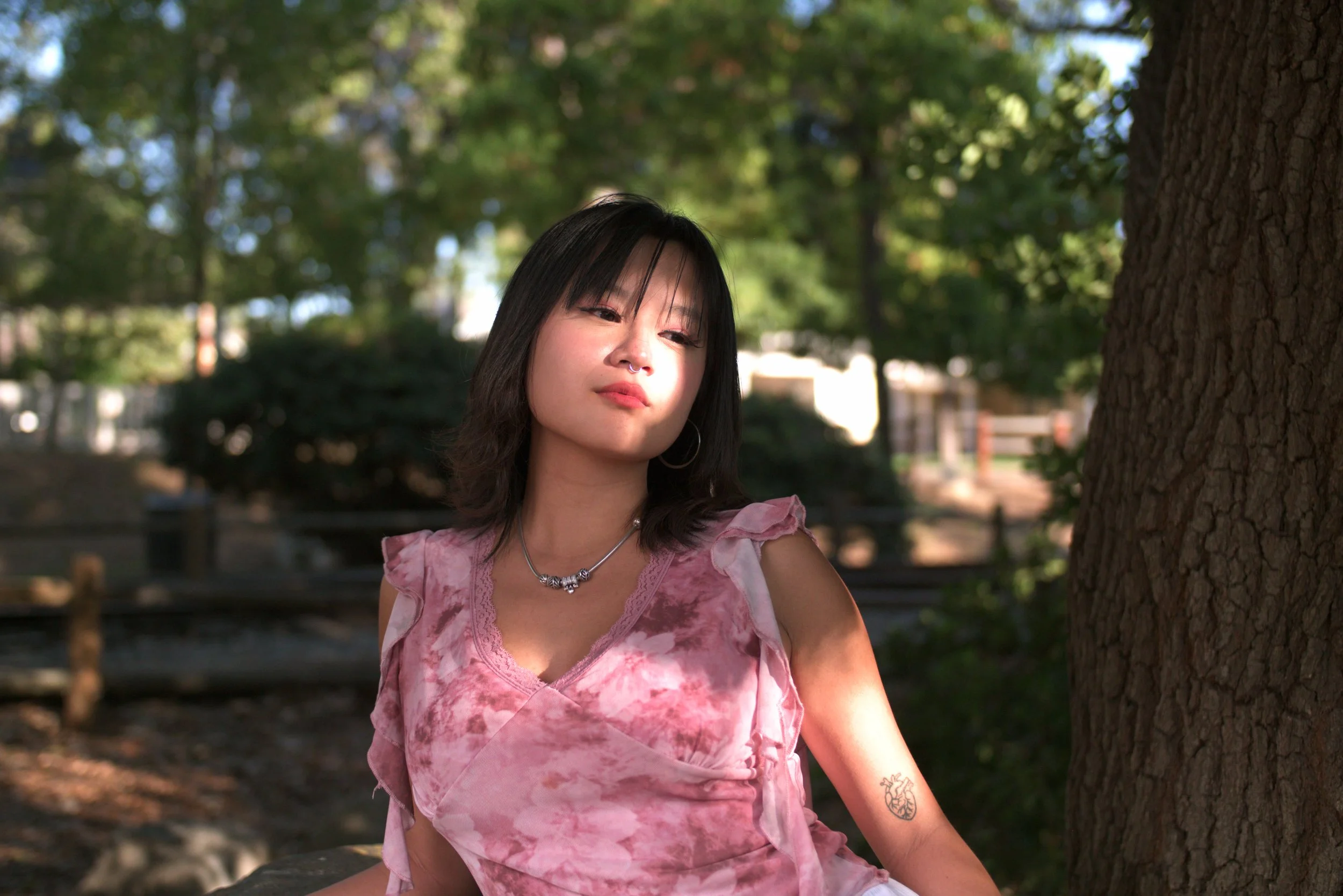 A young woman with black hair and hoop earrings, wearing a pink tie-dye top and a necklace, sitting outdoors beside a tree with sunlight filtering through the leaves.