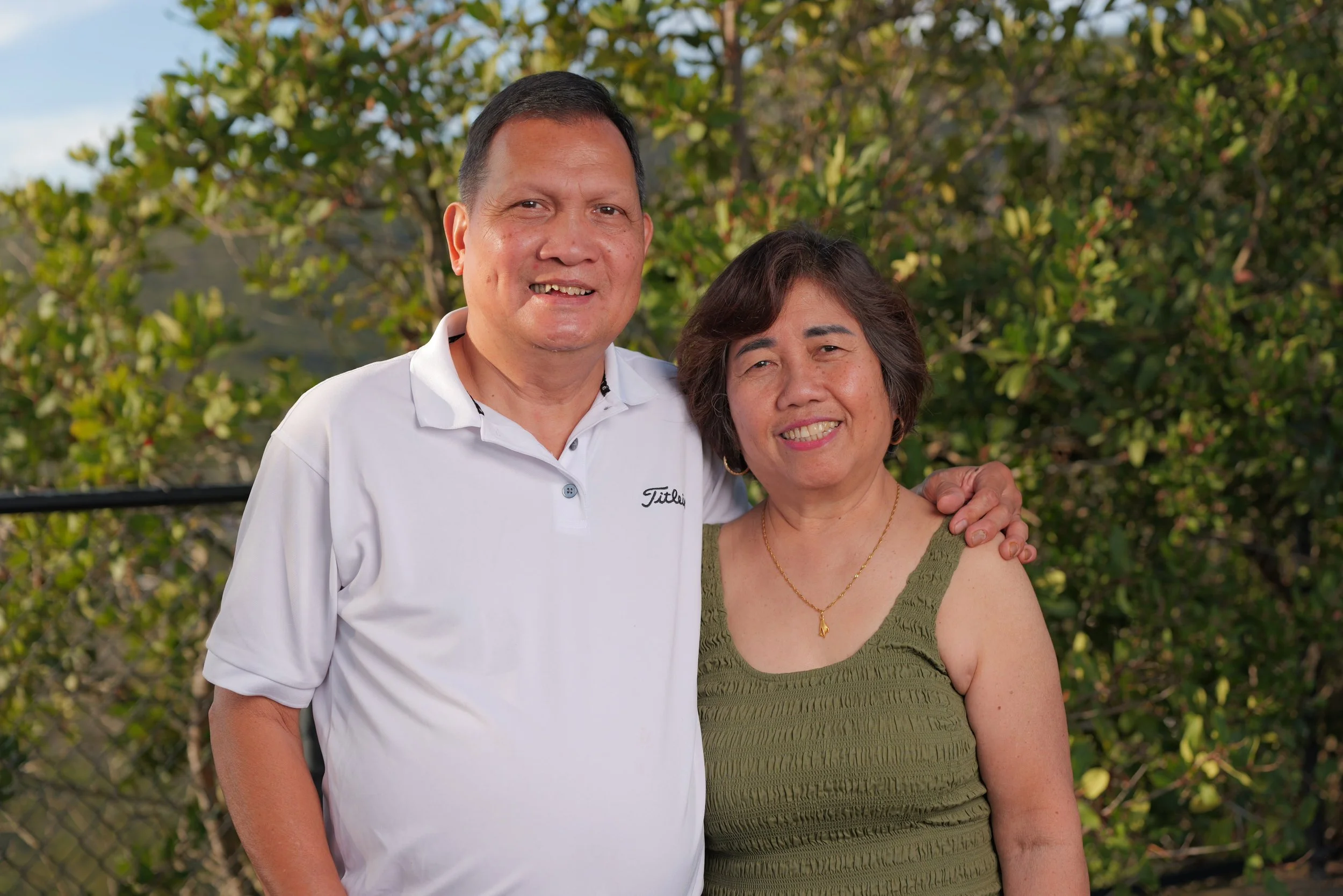 A smiling man and woman standing outdoors, with greenery in the background. The man has short black hair and is wearing a white polo shirt, while the woman has short dark hair and is wearing a green sleeveless top and a gold necklace.