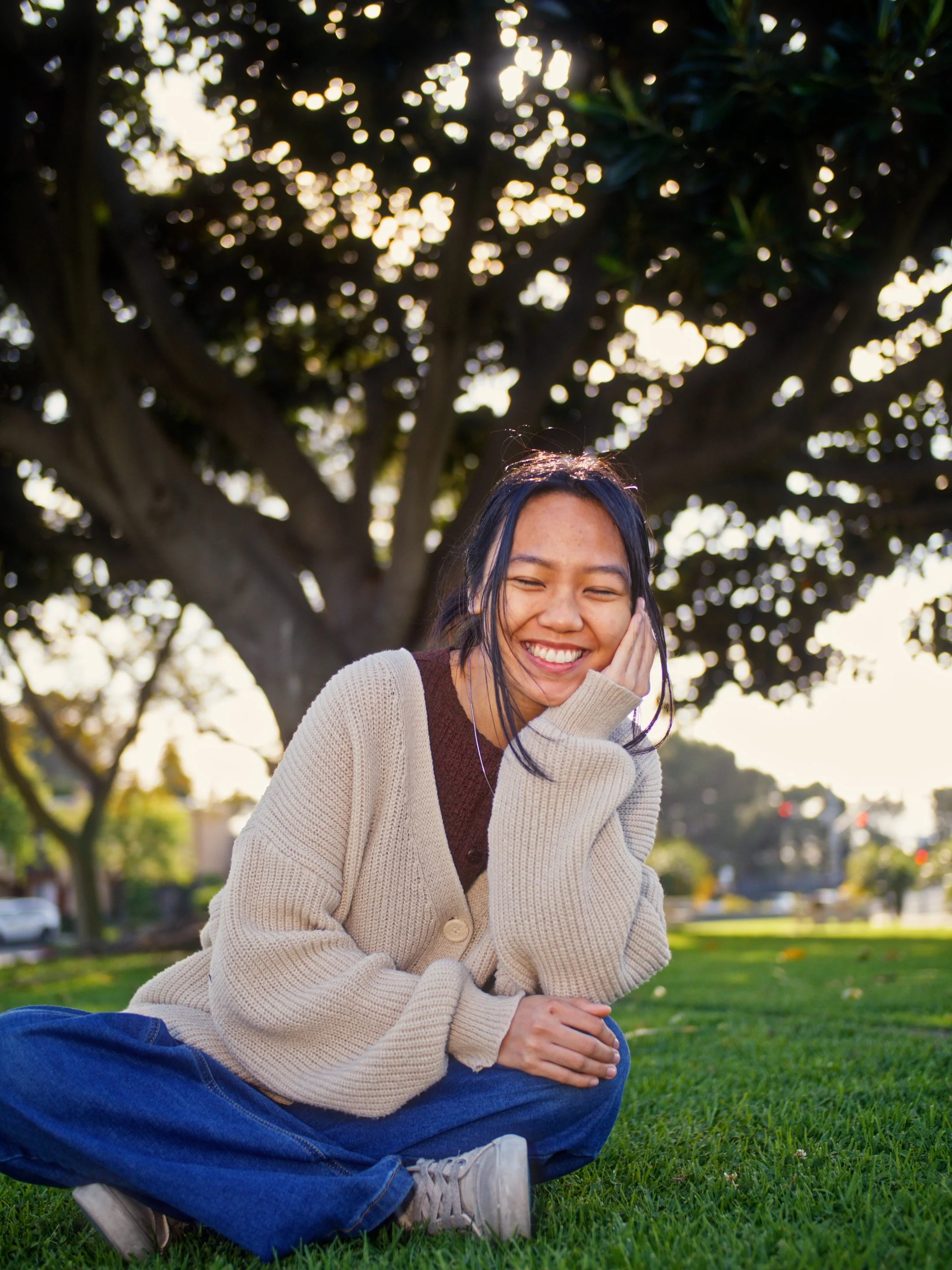 A young woman with dark hair and freckles, smiling and sitting cross-legged on a grassy lawn, with her hand resting on her cheek. She is outdoors near a large tree, with sunlight filtering through the leaves.