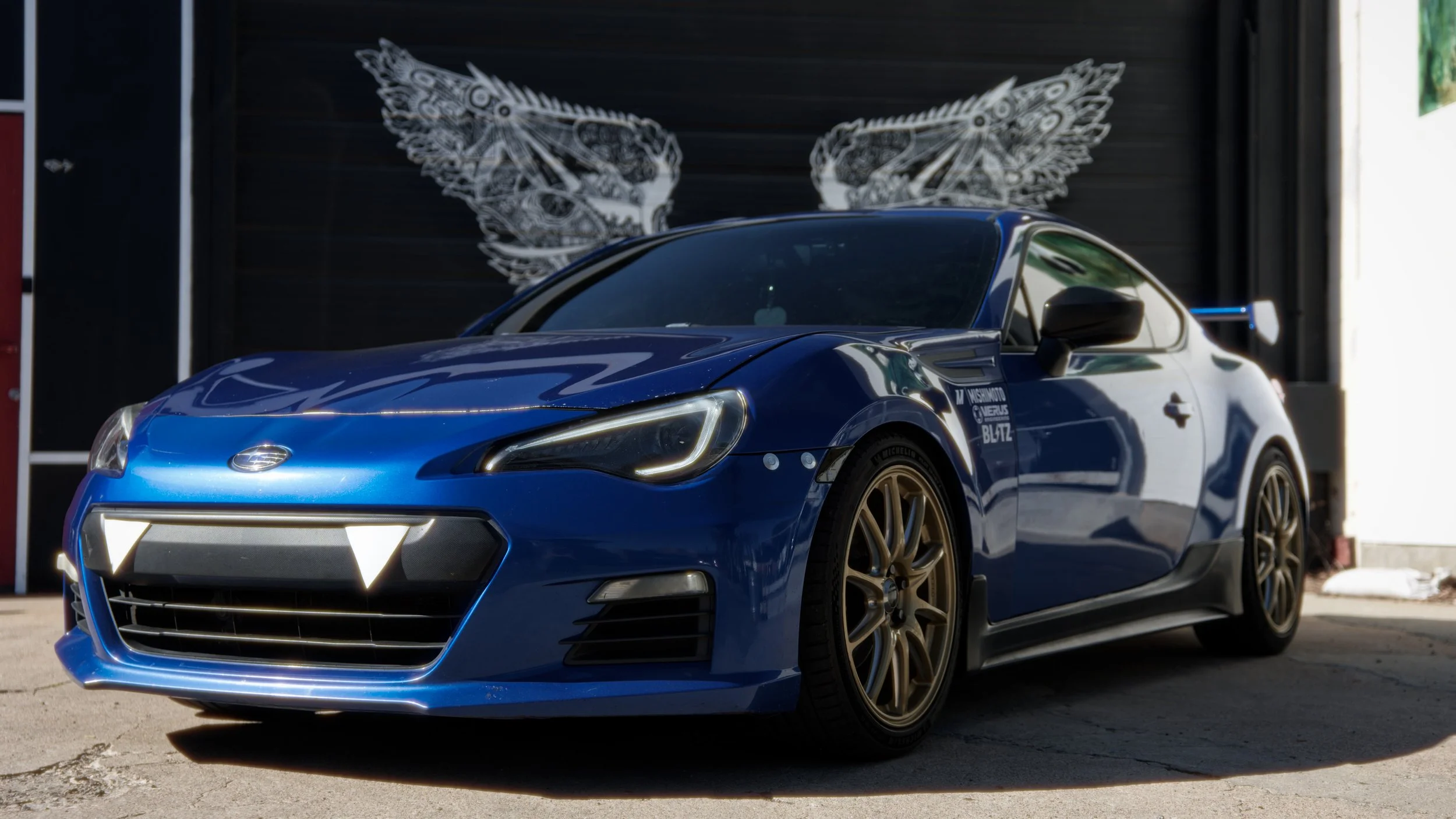 A blue sports car parked outdoors in front of a wall with butterfly wing artwork.