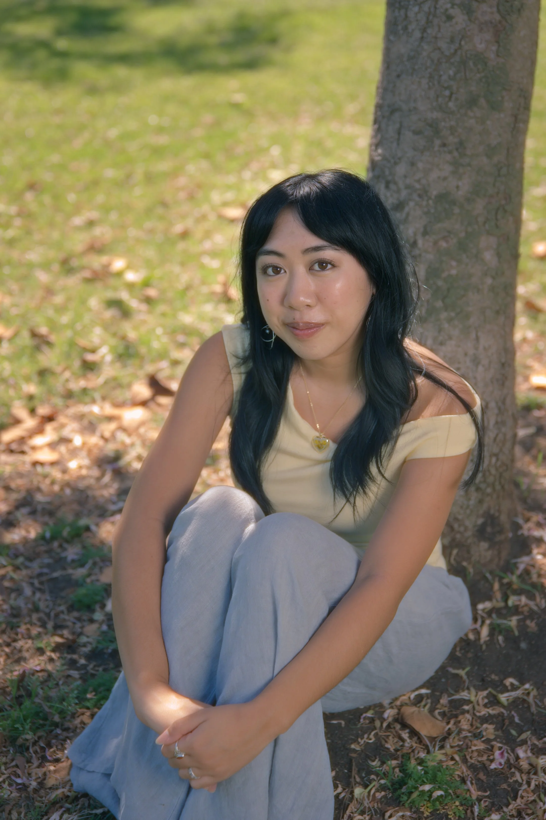 A young woman with black hair sitting on the ground under a tree in a park, wearing a cream-colored top and light-colored pants, with a gentle smile.