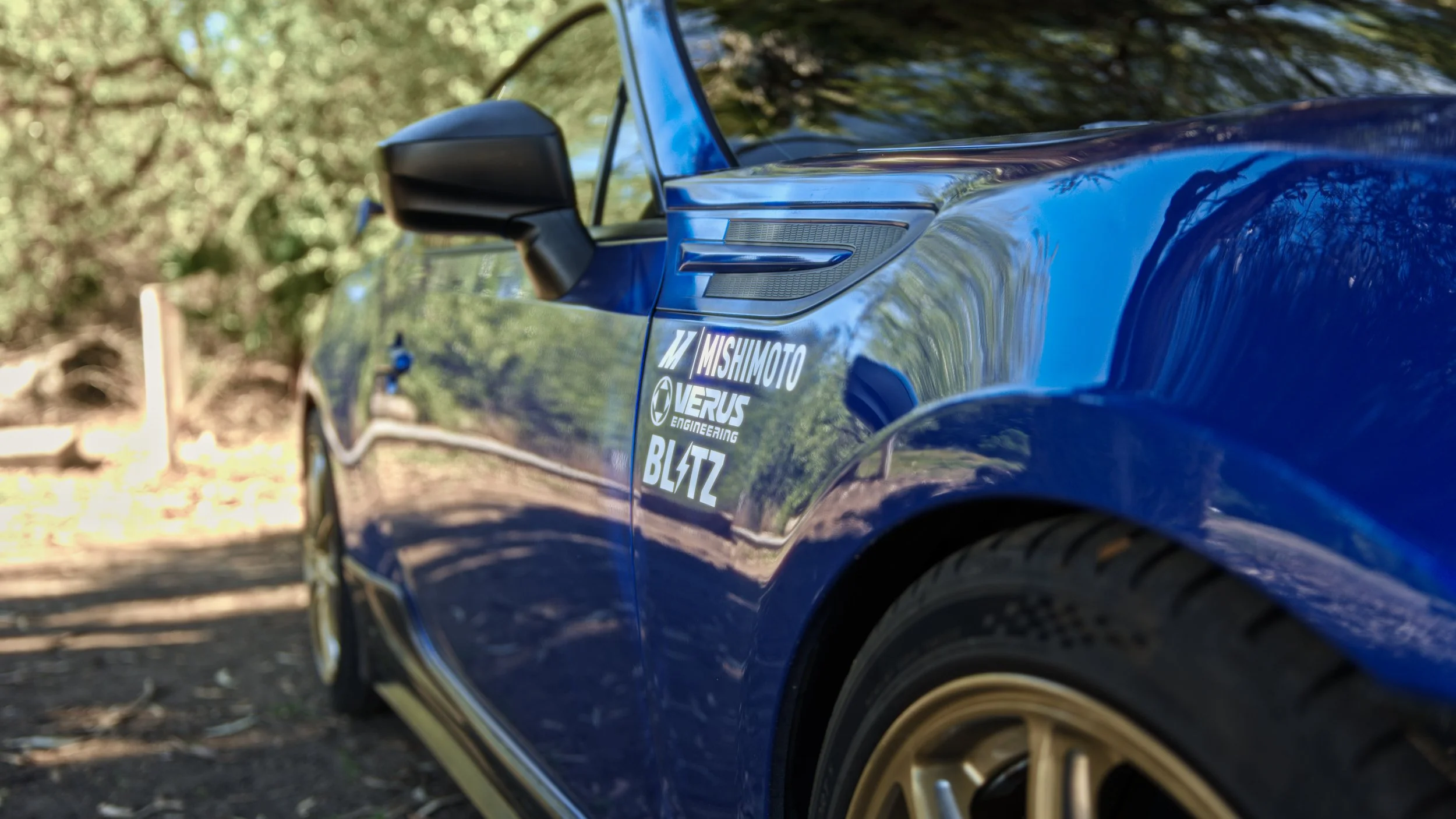Close-up of a shiny blue sports car with sponsor stickers on the side, taken outdoors with trees reflected on its surface.