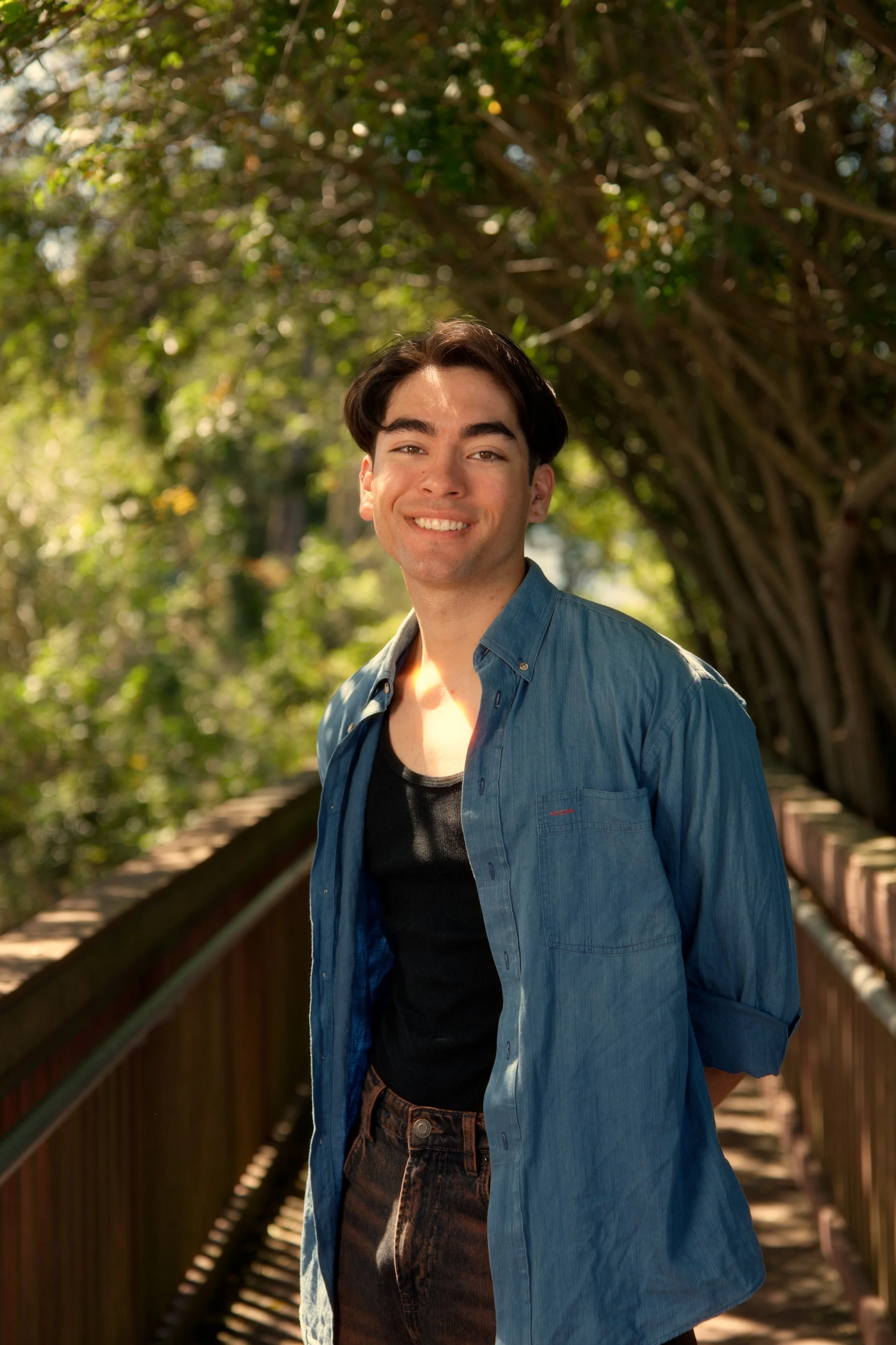 Young man smiling outdoors on a wooden bridge with lush green trees in the background, wearing a dark shirt and a blue button-up shirt.