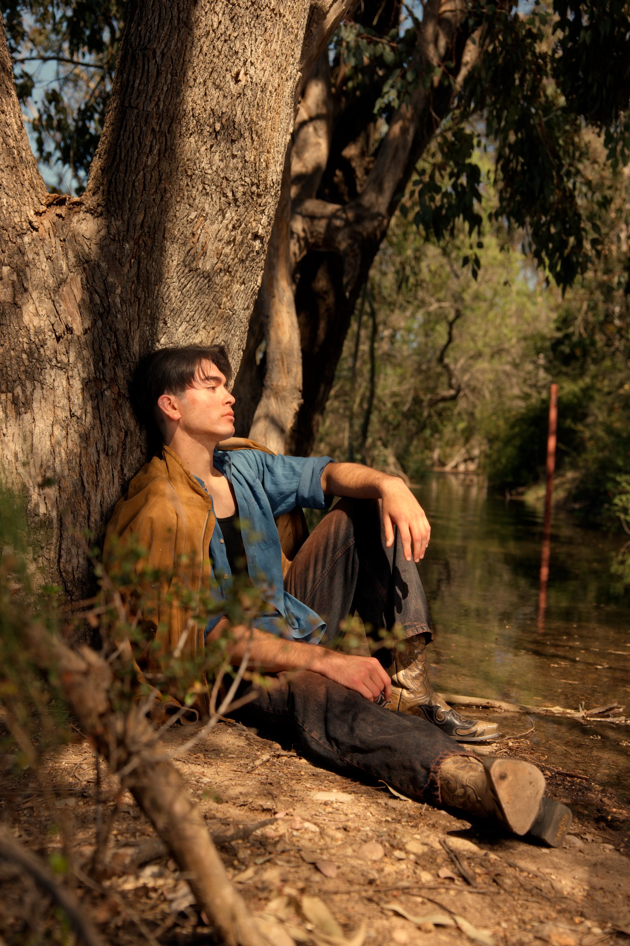 A man sitting with his back against a large tree trunk by a river, with his eyes closed, wearing a blue shirt, brown jacket, and jeans, surrounded by greenery.