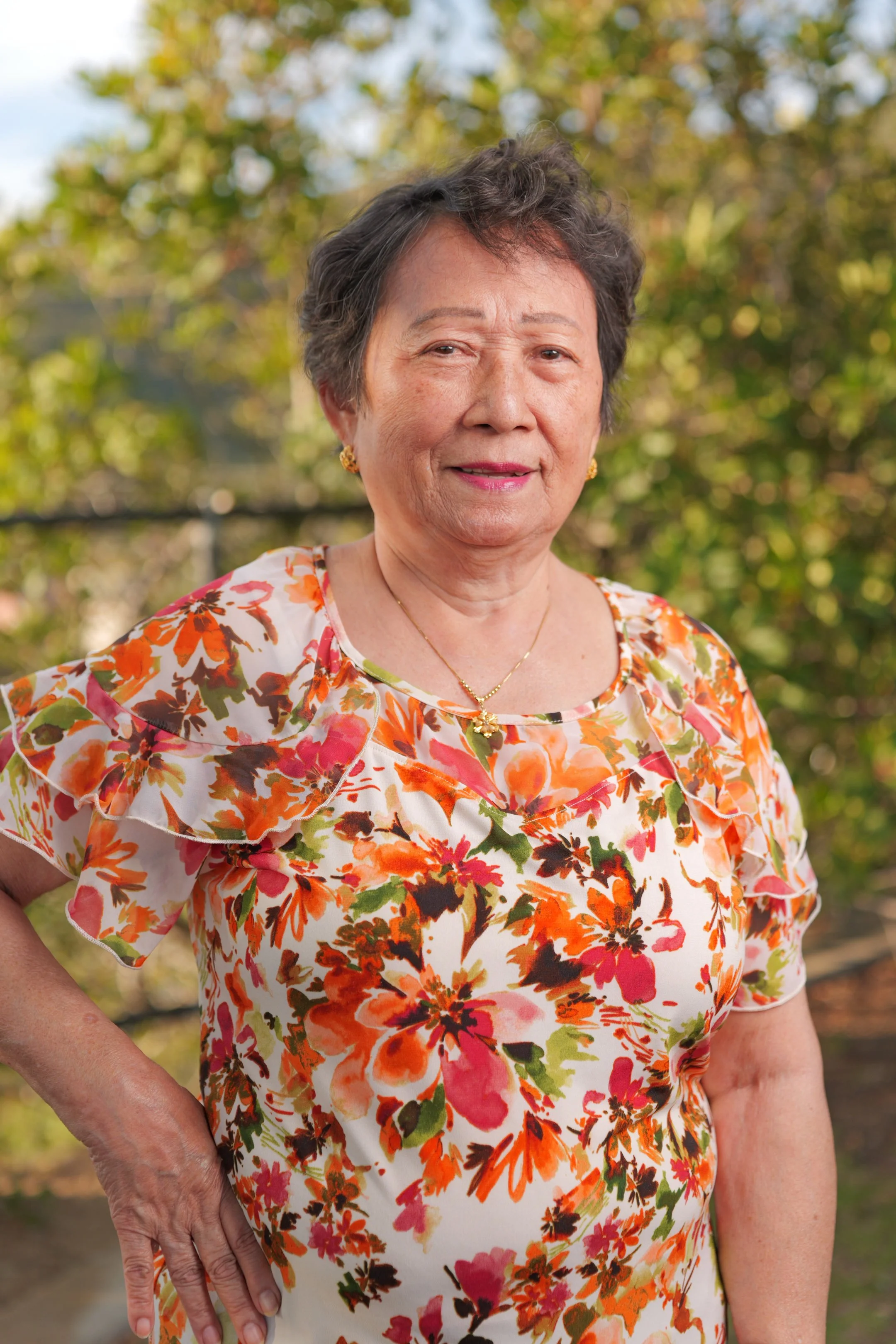 An elderly woman outdoors wearing a floral blouse with orange, pink, and green colors, standing with her hand on her hip and a slight smile, with trees and a fence in the background.