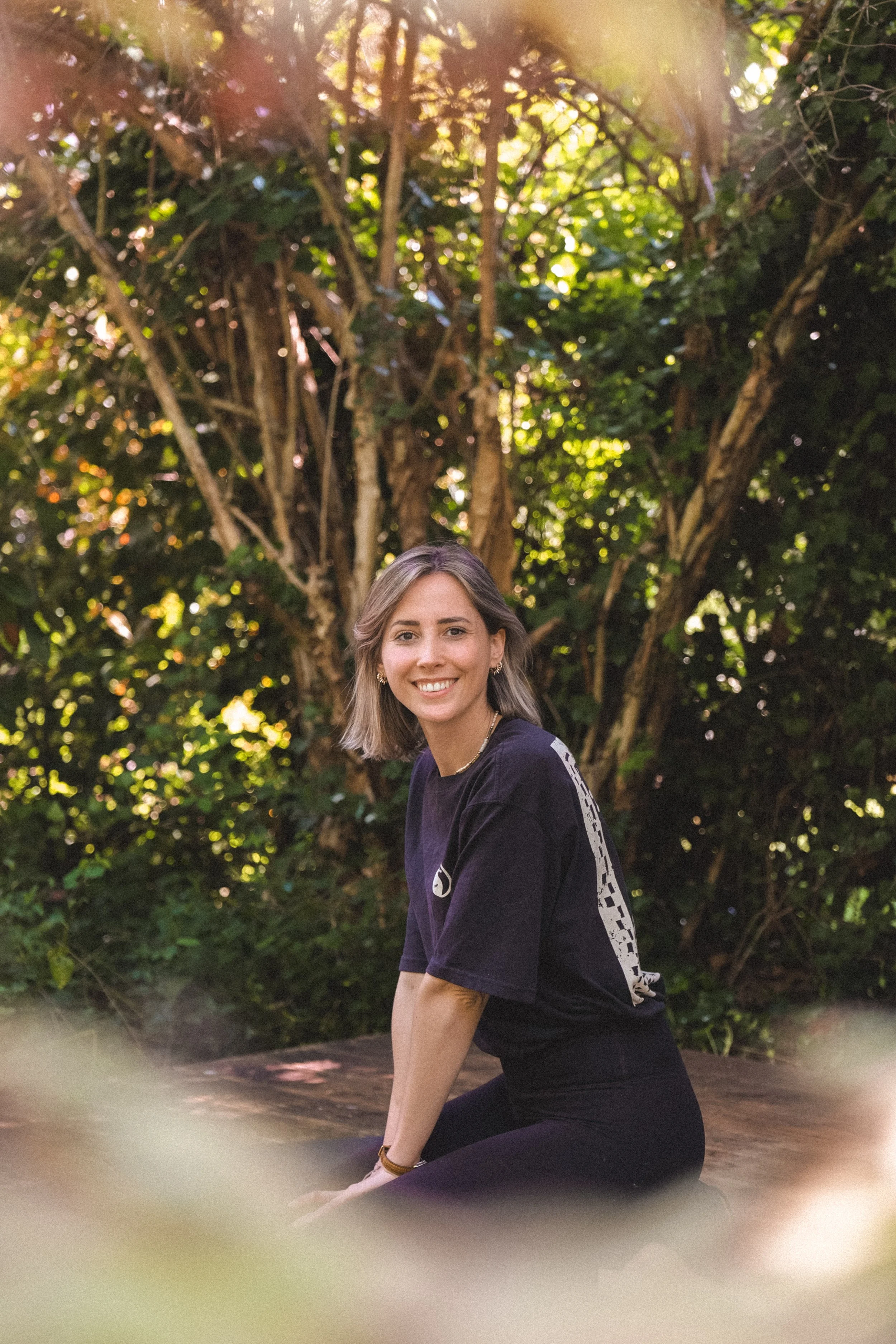 Junge Frau in dunklem T-Shirt beim Sitzen im Freien vor einem Baum mit grünen Blättern an einem sonnigen Tag.