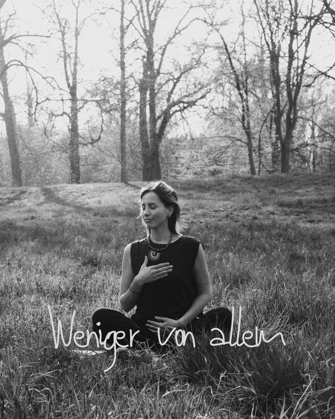 Schwarz-Weiß-Foto einer jungen Frau, die im Sitzen im Gras in einem Wald sitzt, mit geschlossenen Augen und Hand auf der Brust. Text auf dem Bild: "Weniger Von allem".