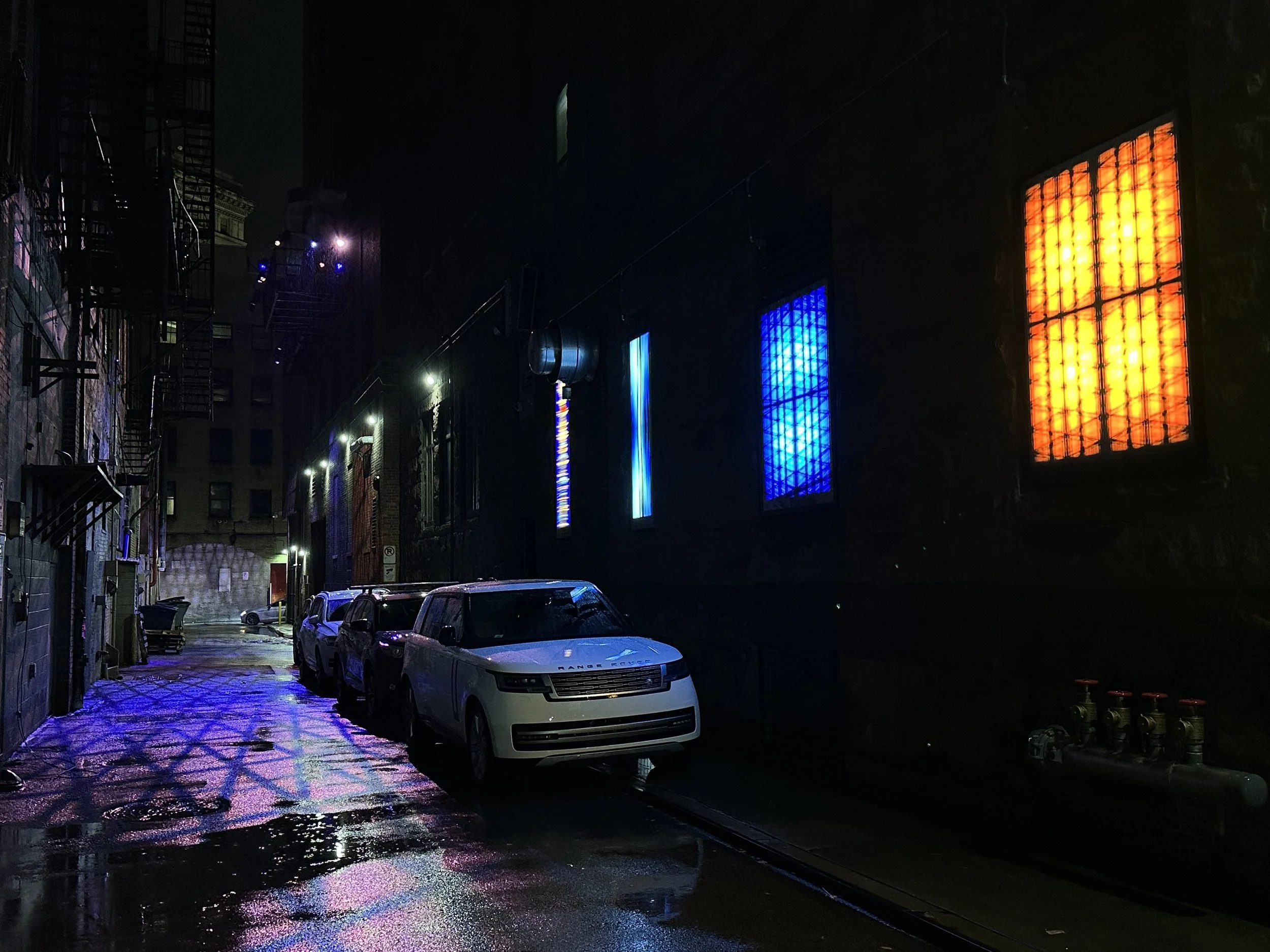 Dark alley with wet pavement reflecting colorful neon lights on surrounding buildings and a few parked cars.
