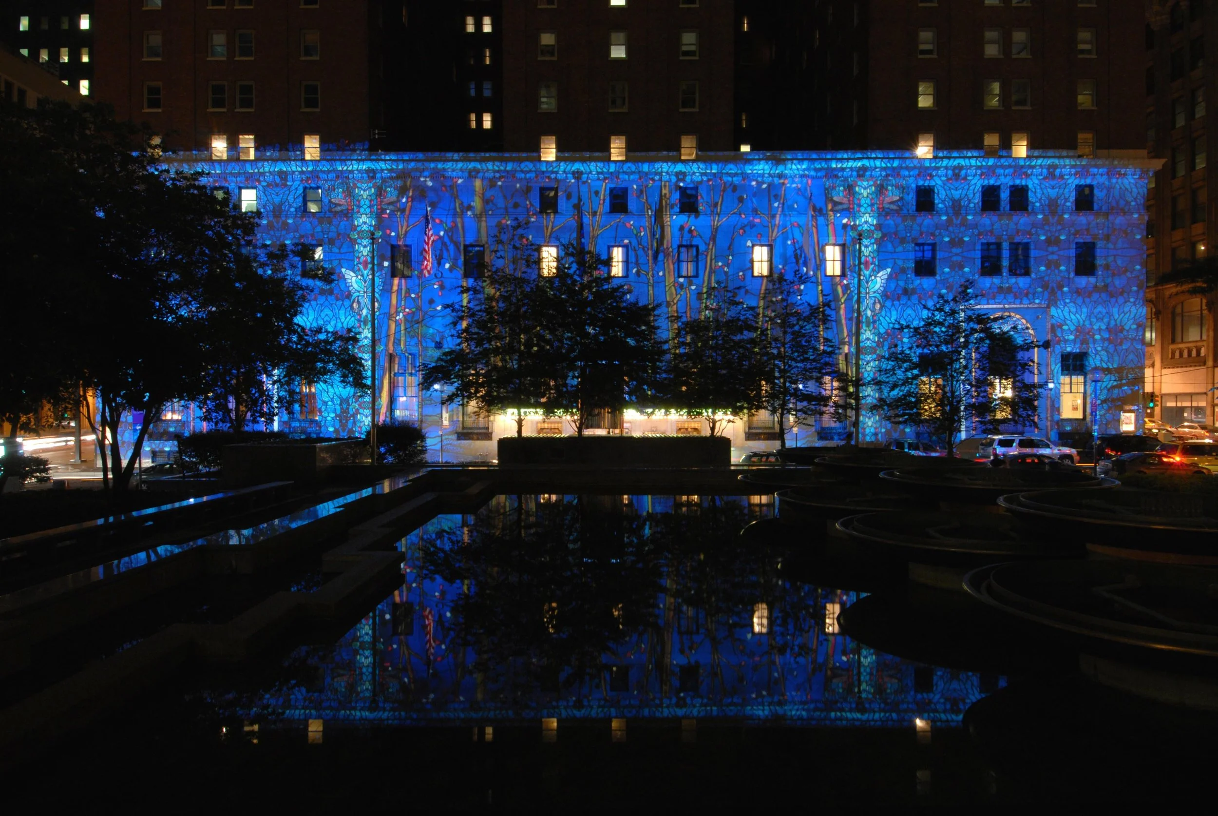 Building illuminated with blue and colorful light projection at night, trees and parked cars in foreground, reflection in water.