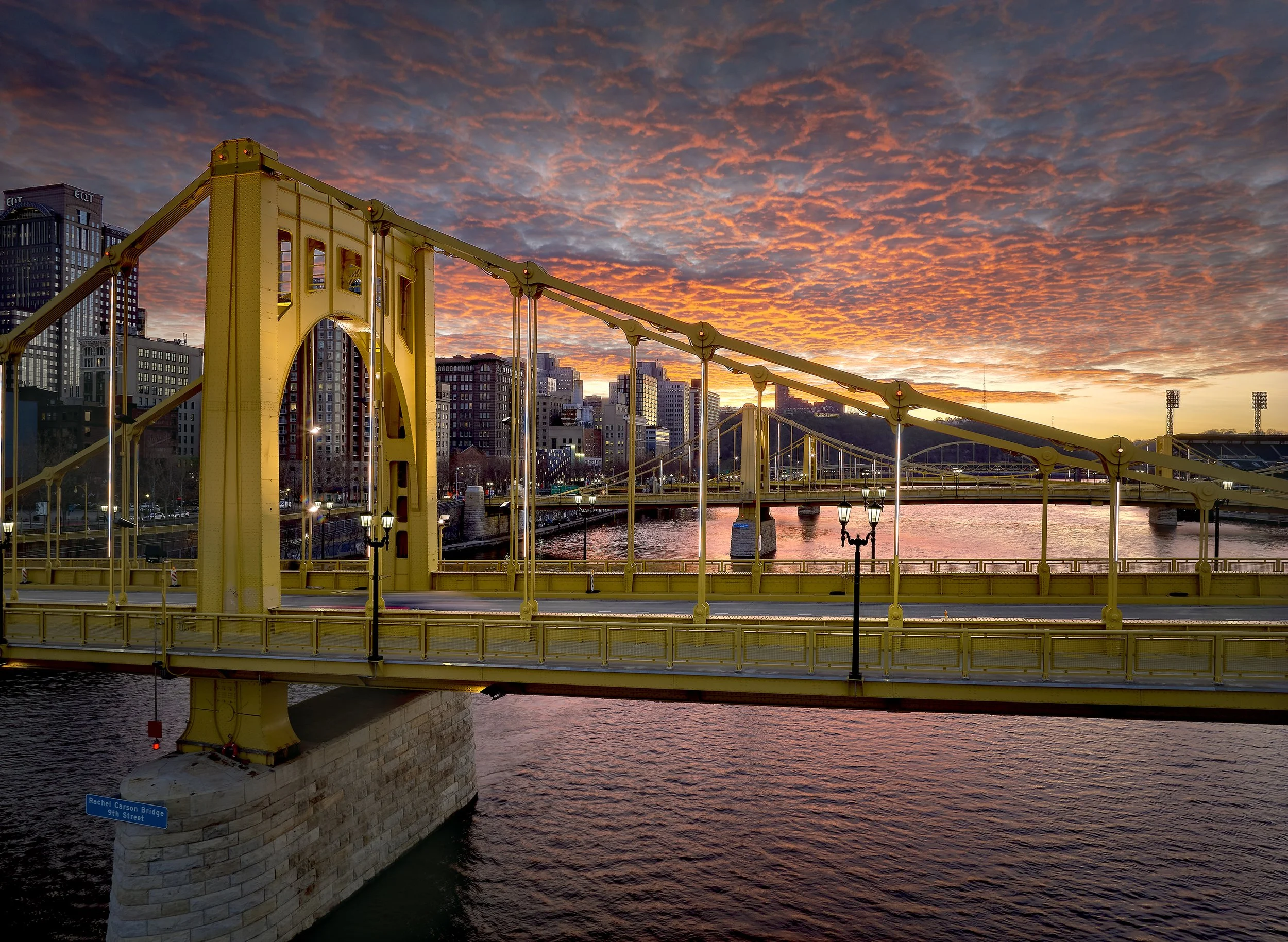 Sunset over a city skyline with a yellow suspension bridge crossing a river in the foreground.