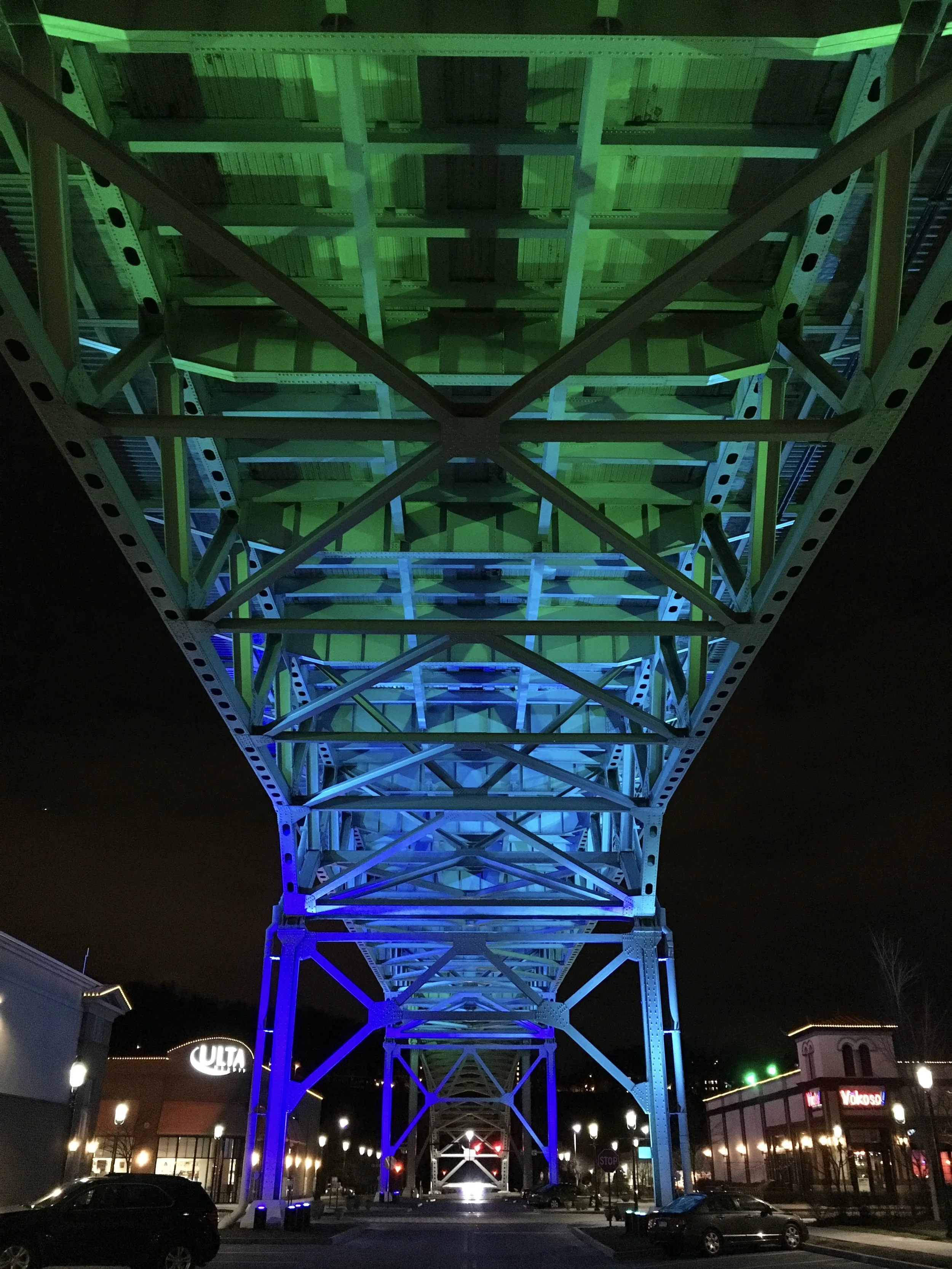 Night view of a lit-up observation tower with colorful blue and green lighting, situated in a shopping area with cars parked below and illuminated signs for stores such as ULTA Beauty and Yokoso.
