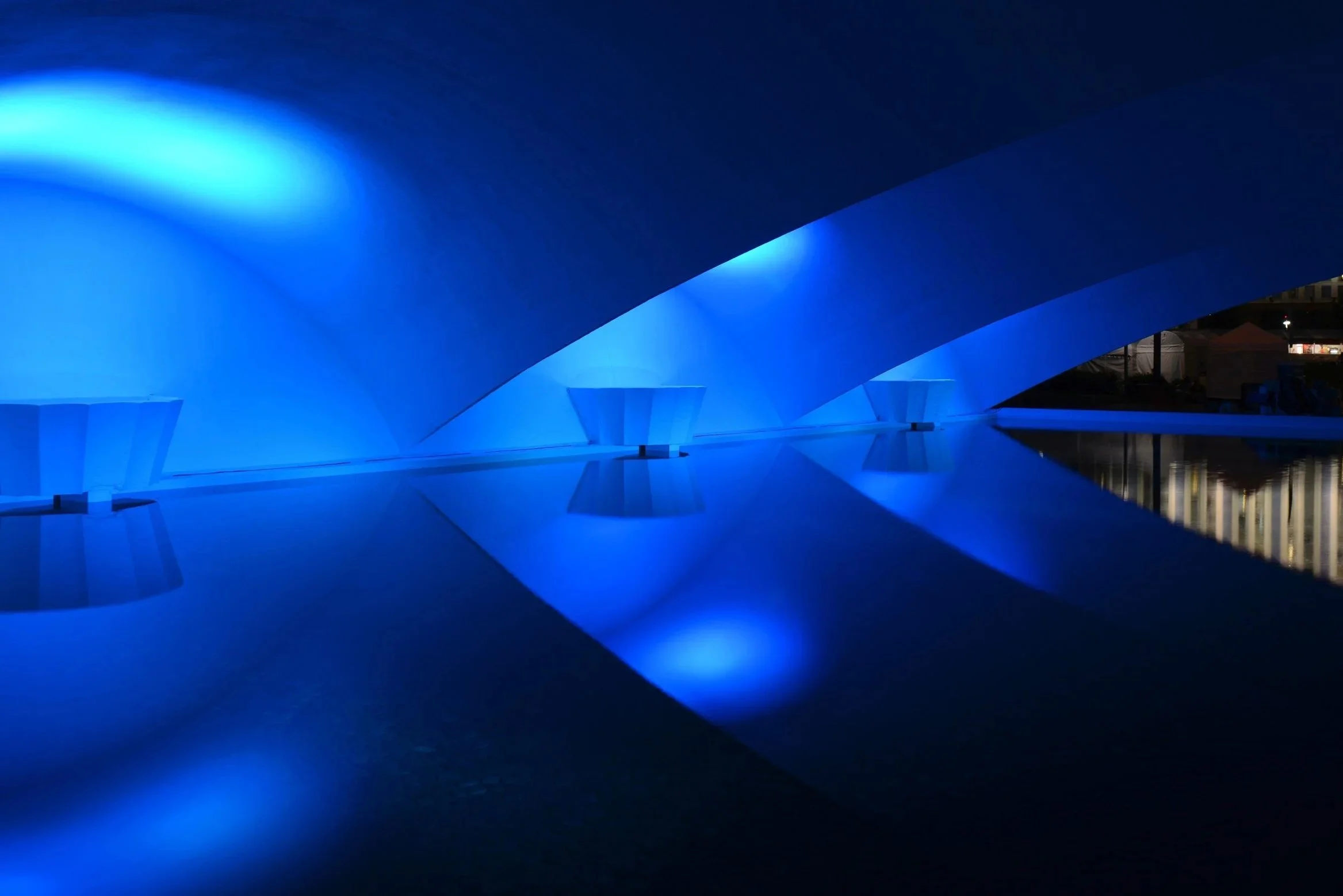 A nighttime view of a modern architectural structure illuminated in blue, with curved and angular surfaces and reflecting pool.