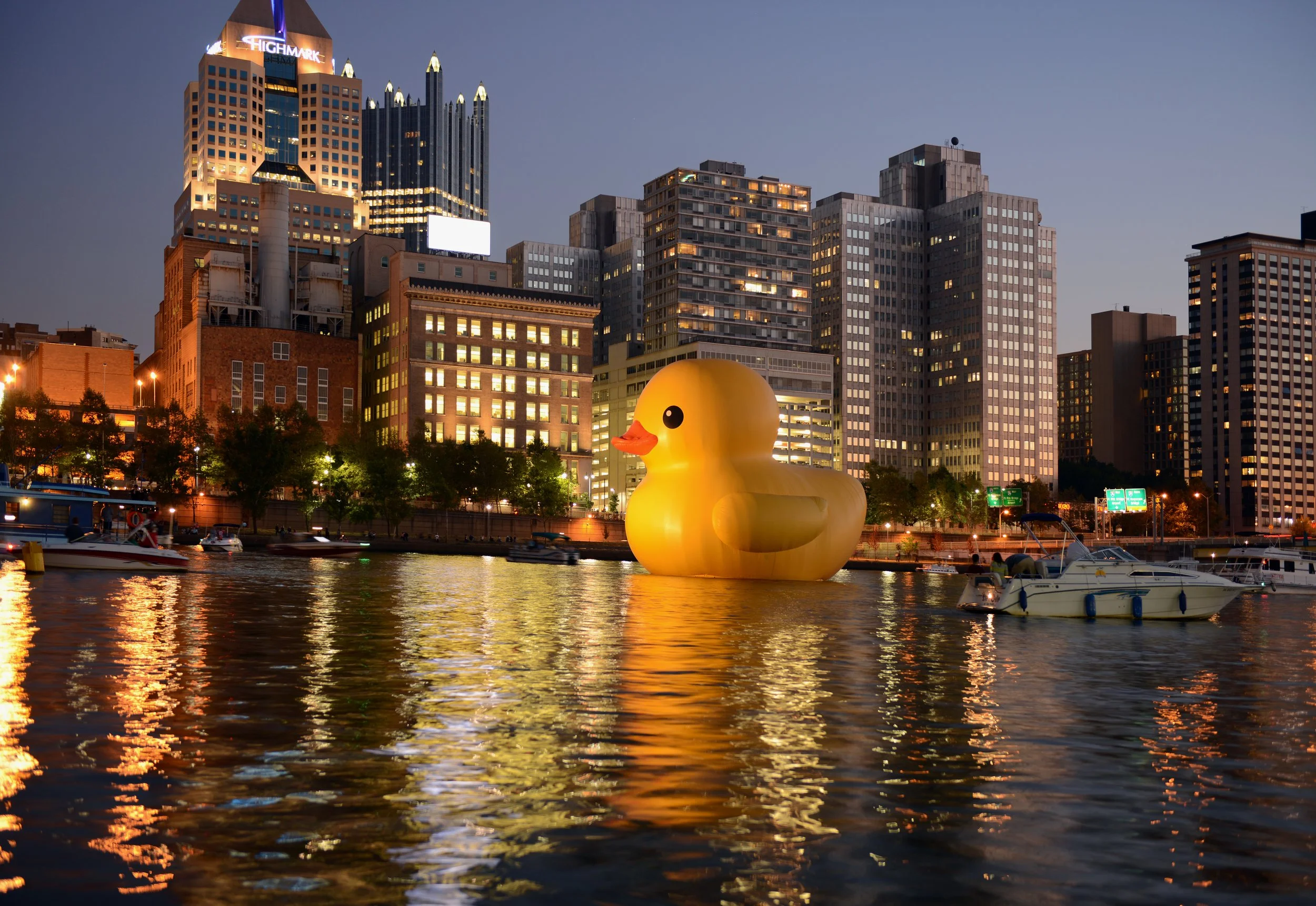 A large yellow rubber duck floating on a river with city high-rise buildings in the background at dusk.
