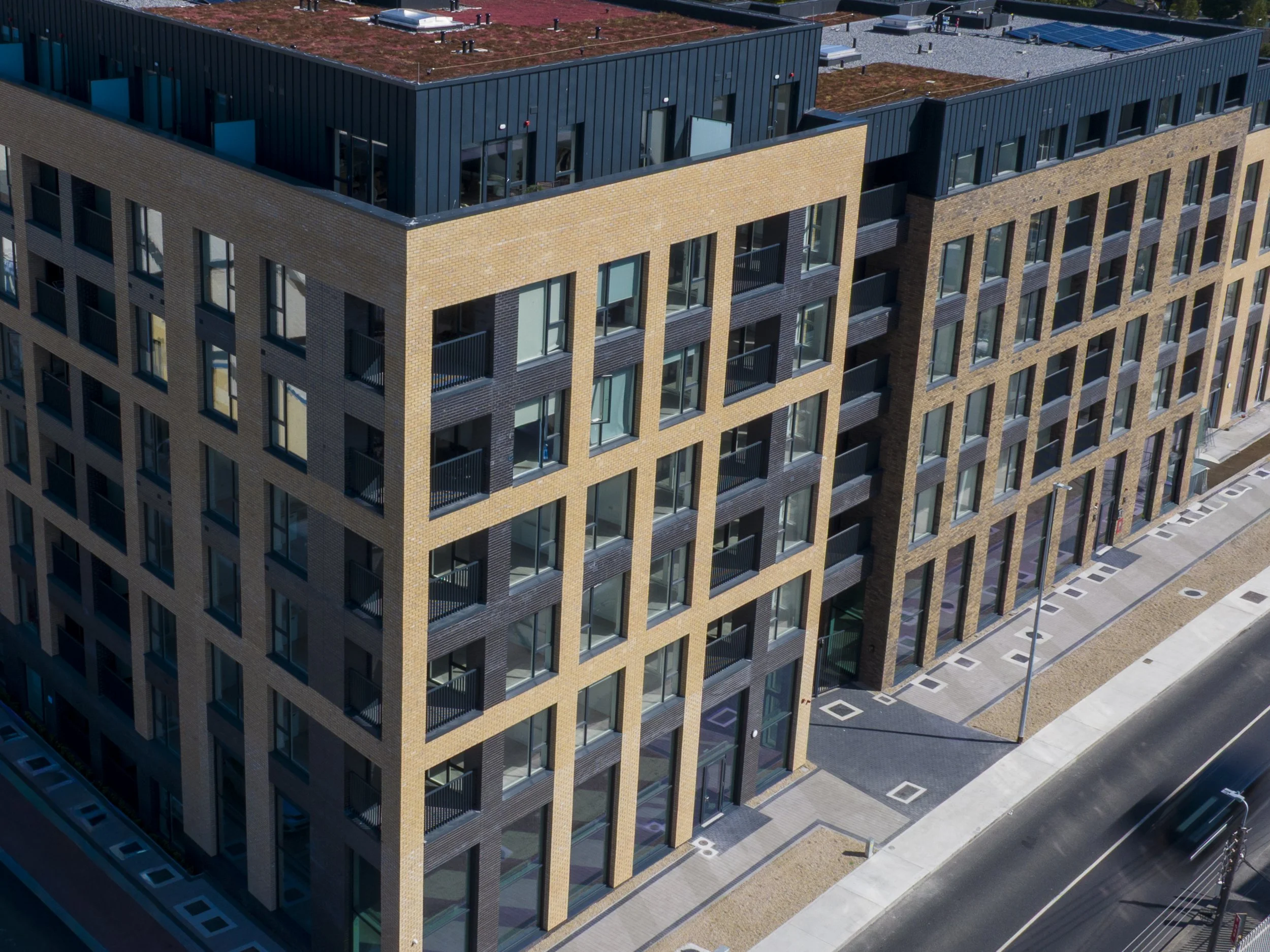An aerial view of a modern multi-story apartment building with brick and blue panel exterior, multiple glass doors and windows, balconies, and a surrounding sidewalk and street.