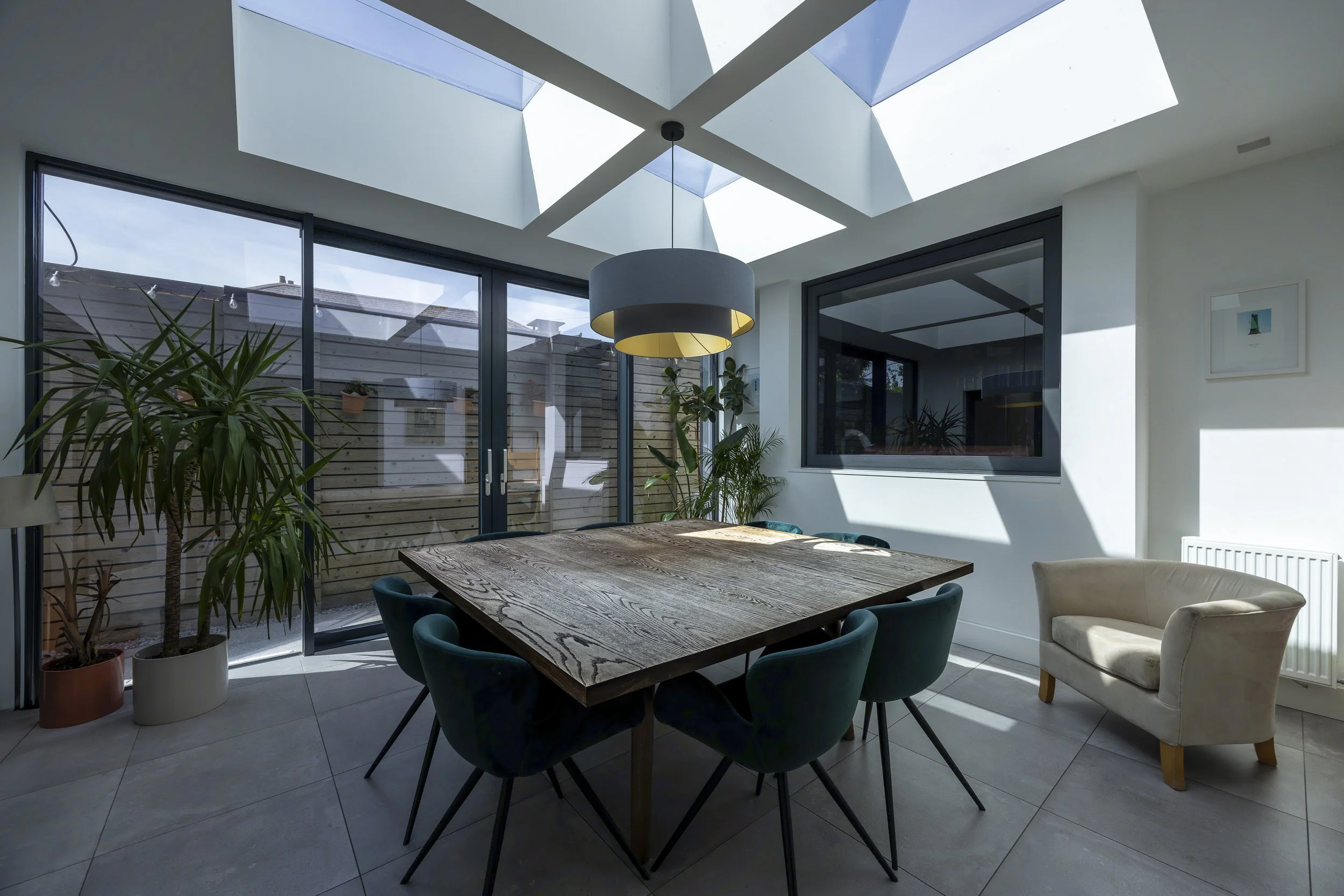 A sunlit dining room with large skylights, a wooden dining table with six teal chairs, an armchair, potted plants, and a sliding glass door leading to a fenced outdoor space.