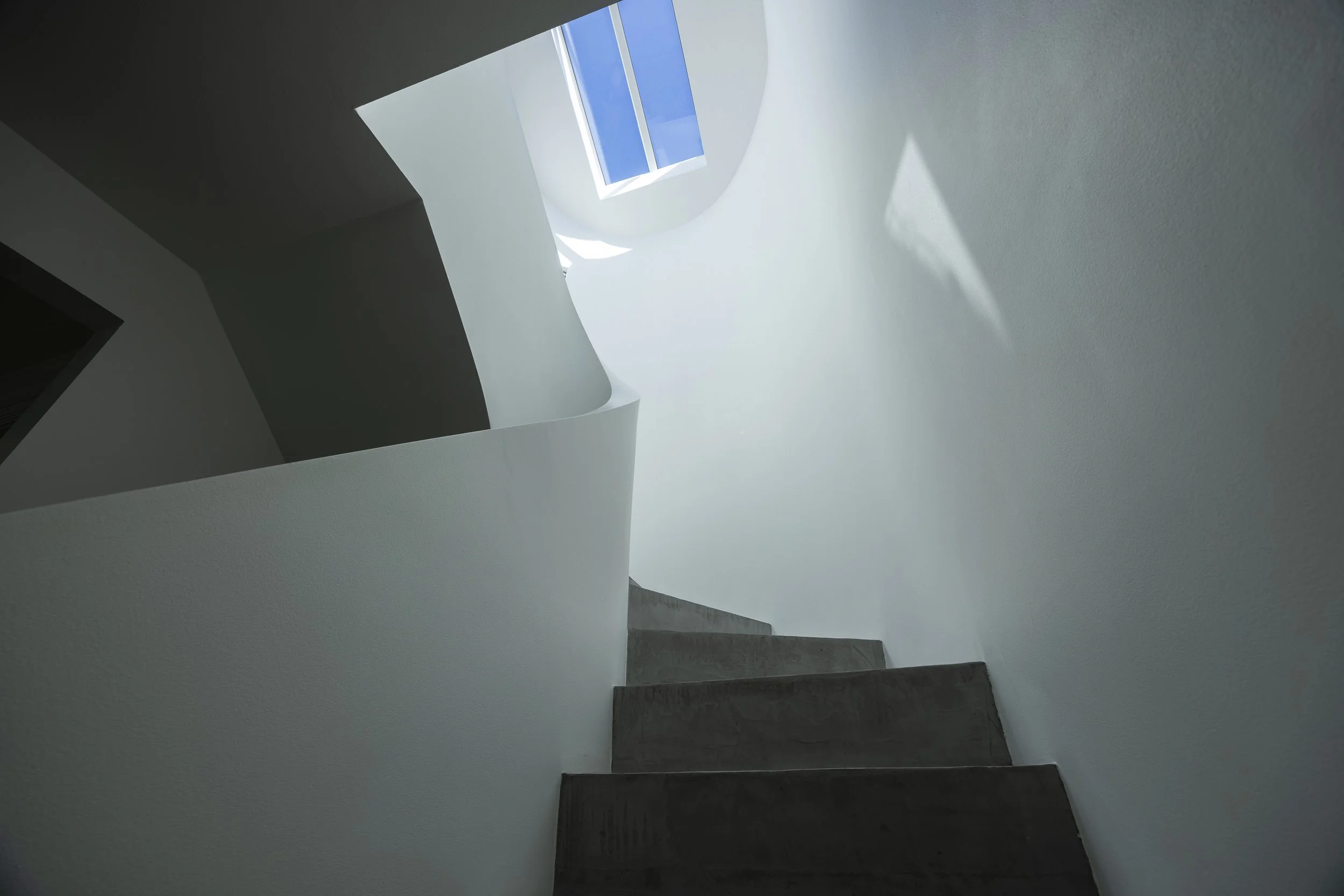 View of a modern staircase with dark steps and white walls, looking up toward a skylight with sunlight and blue sky visible.